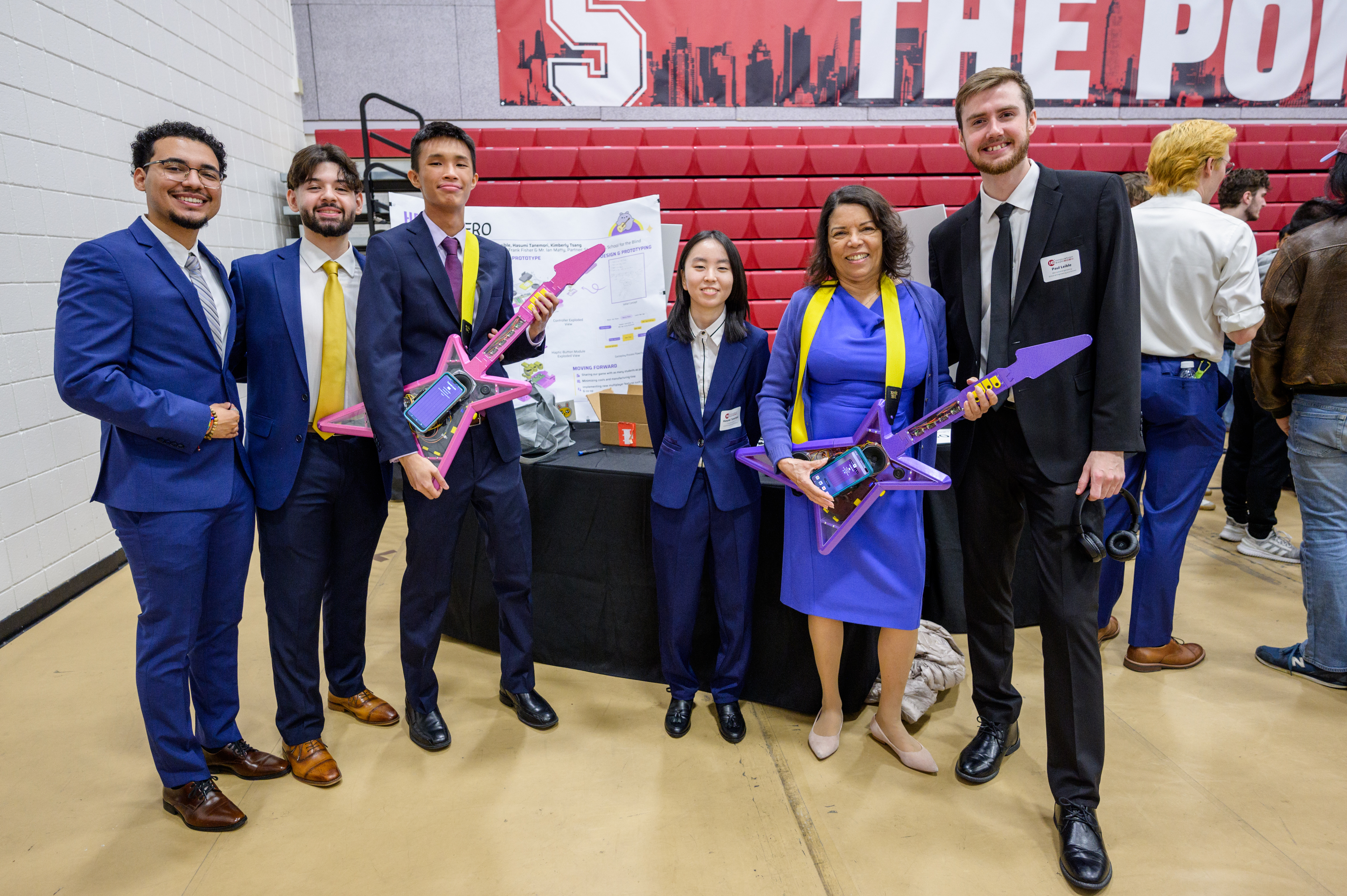 A student on the left plays a purple electric guitar and is guided by a teacher who helps her press the correct guitar buttons of  the Haptic Hero game. 