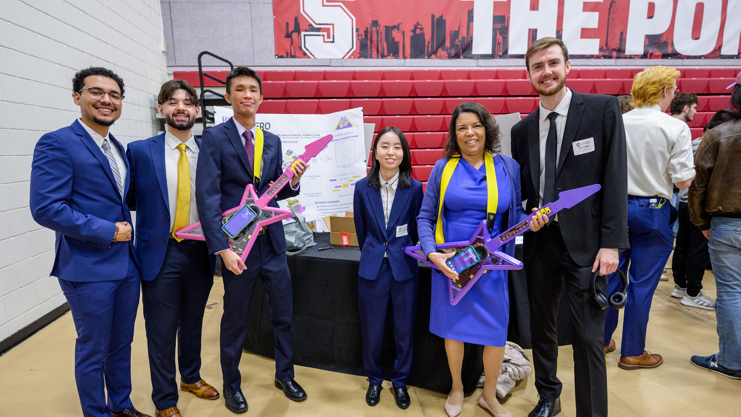 A student on the left plays a purple electric guitar and is guided by a teacher who helps her press the correct guitar buttons of the Haptic Hero game.