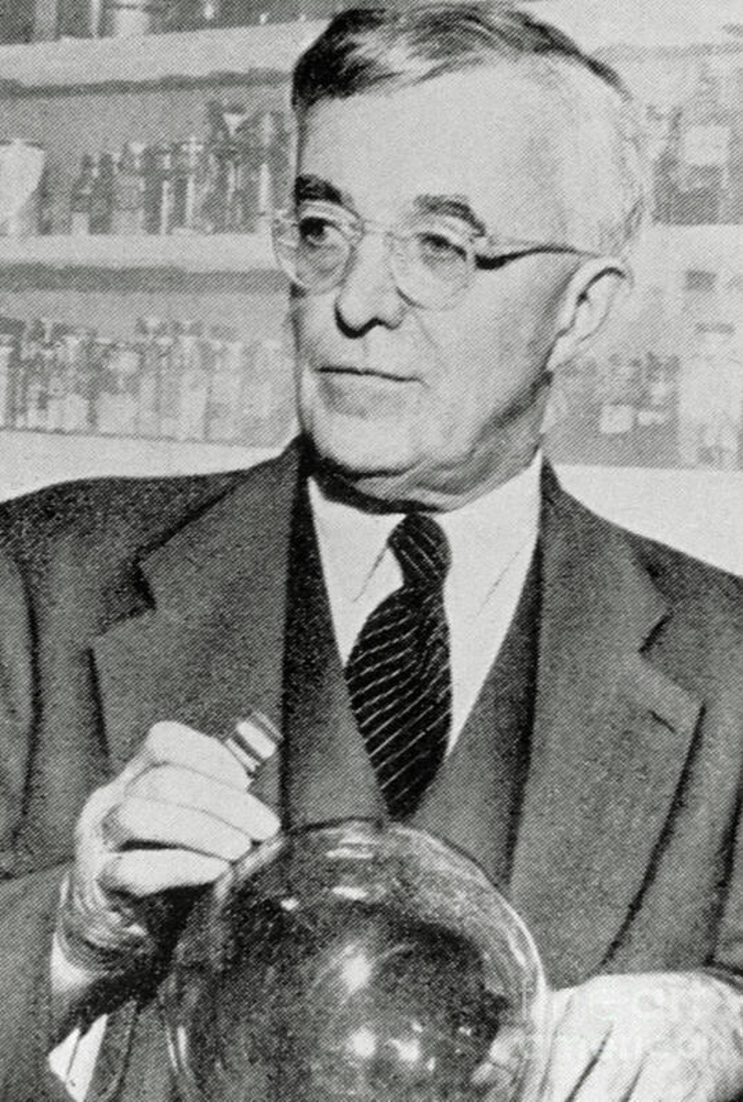 A white male in a dark suit and glasses looking to his right with a circular globe-like glass object in his hands.