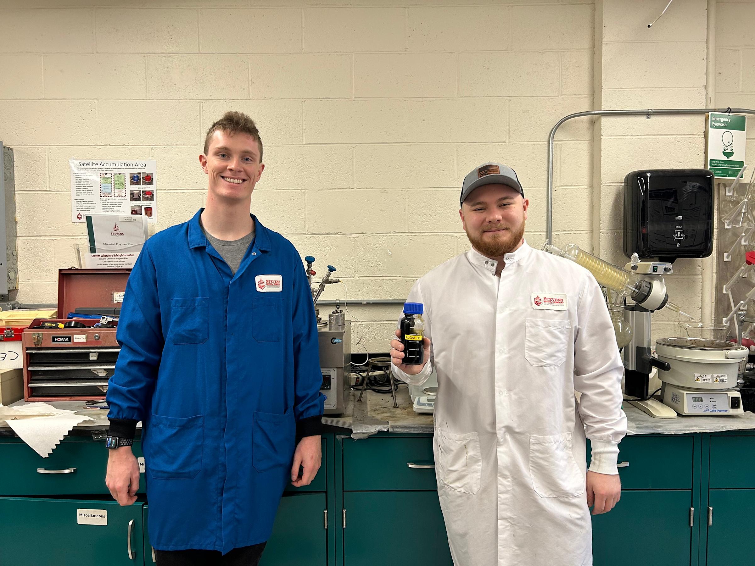 Two students in blue and white lab coats stand in a lab. The student on the right holds up a bottle labeled "Algal Oil."