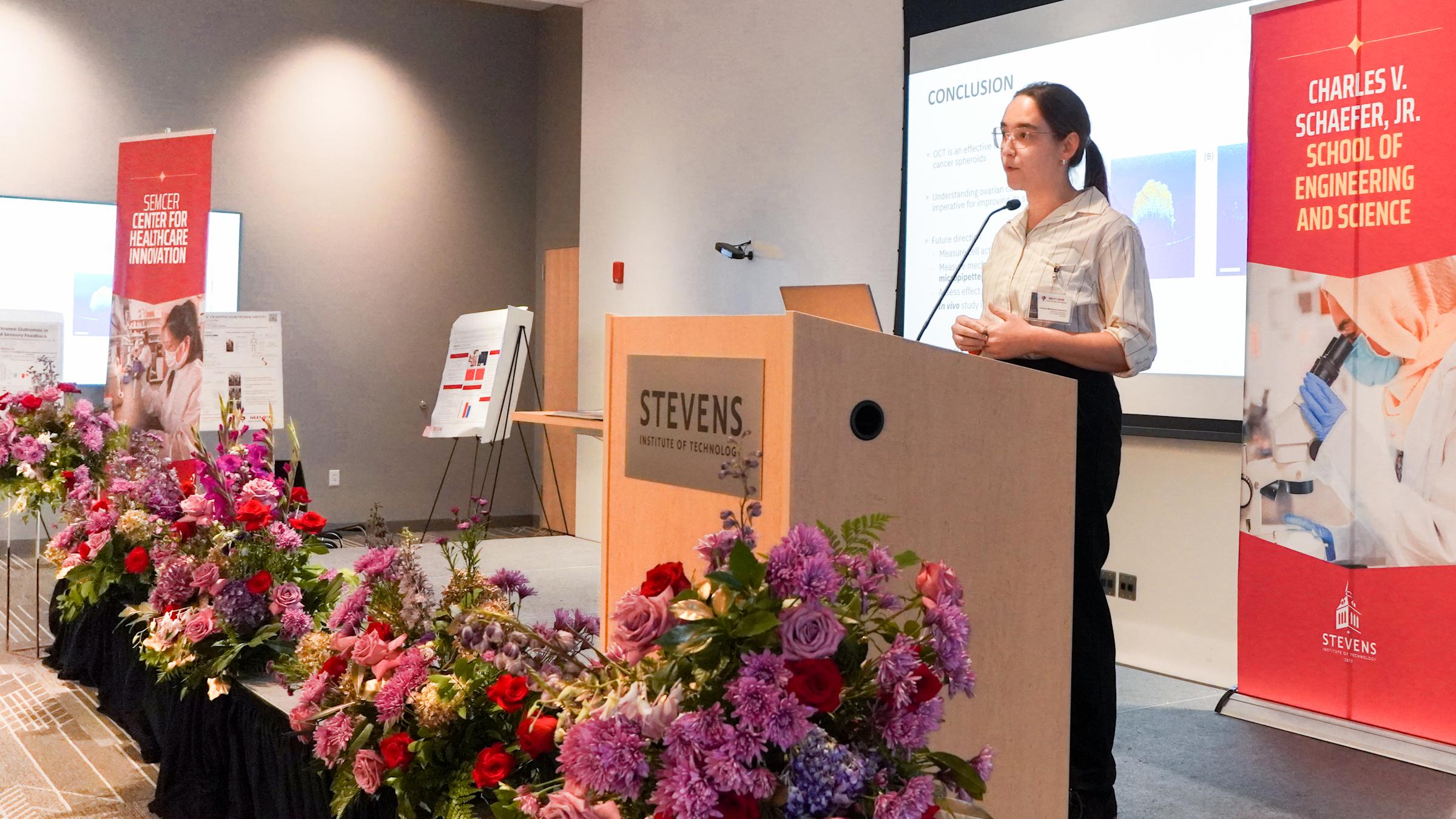A student presents on the stage at the Next Gen Healthcare Innovators Symposium 2024 with a backdrop of research slides in front of flowers.