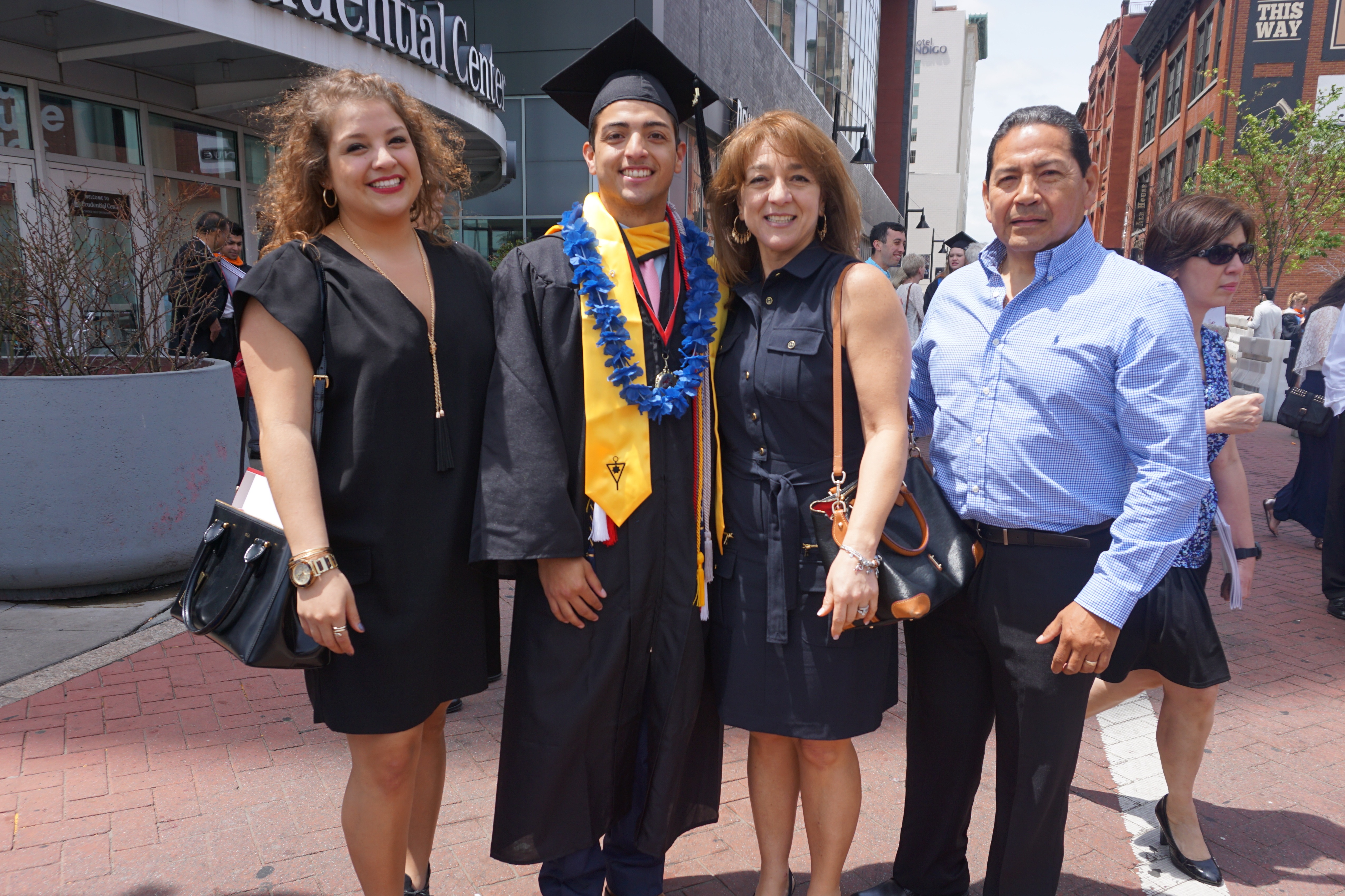 Four people on a sidewalk after a graduation ceremony. A young man in a black cap and gown with blue and gold honor cords and stoles stands between two women in dark dresses and an older man in a light blue checked shirt, with tall buildings behind them.
