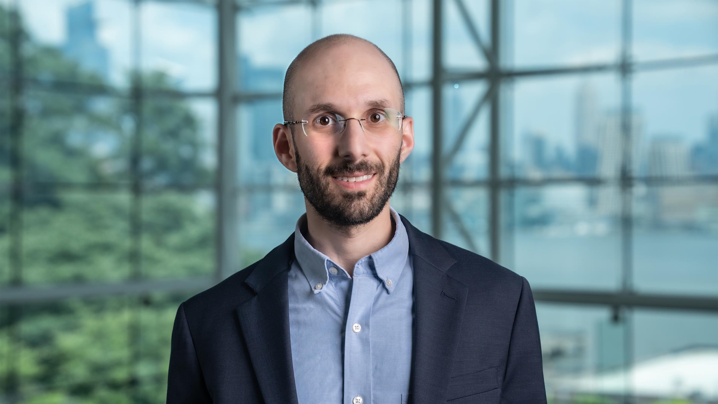 Zach Feinstein stands in the Babbio Center with the New York City skyline visible in the background through the windows.