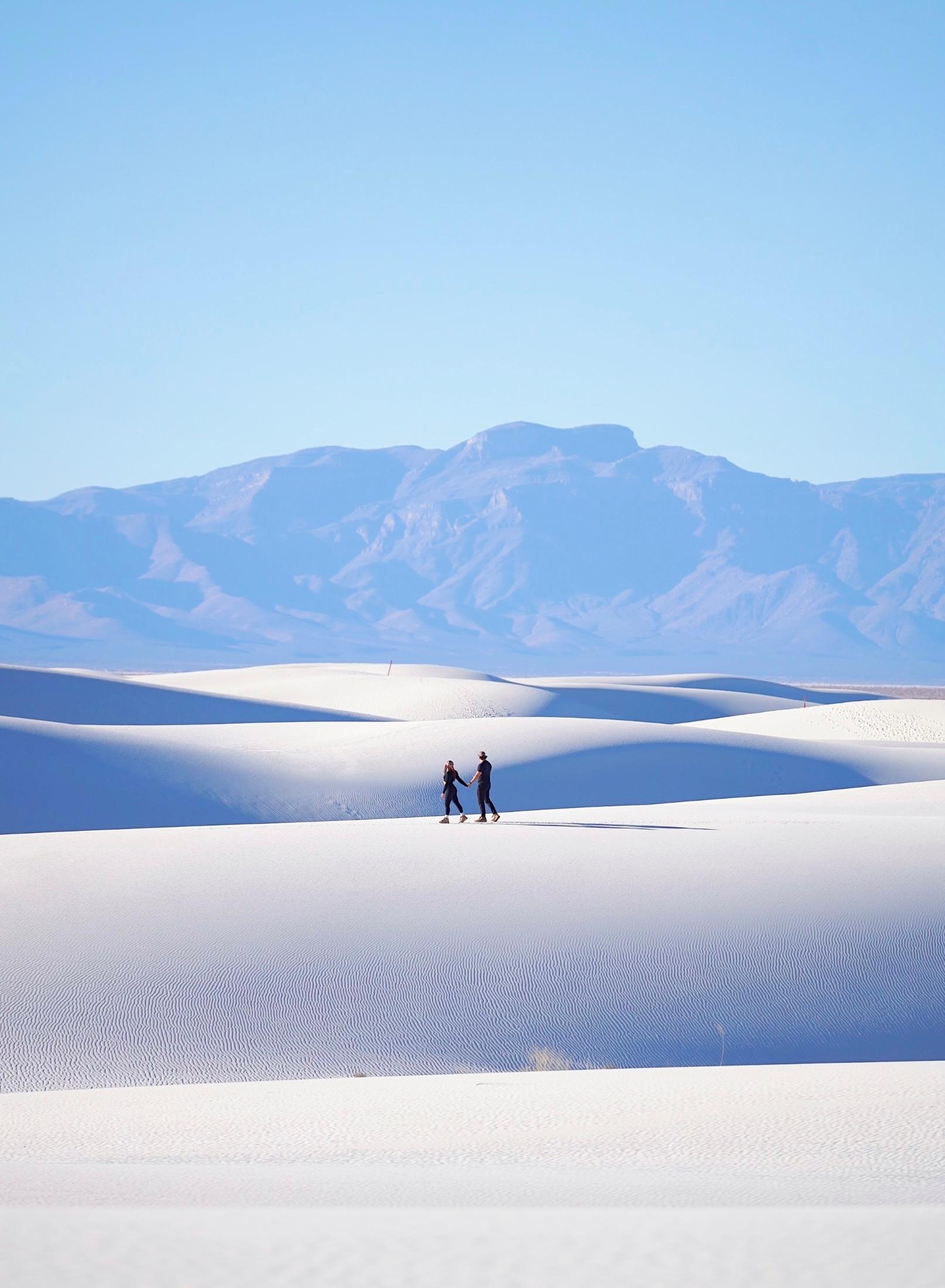 Two people hold hands while walking across vast white gypsum sand dunes, appearing small against the sweeping landscape. Blue mountain ranges stretch across the horizon under a clear sky, likely White Sands National Park, New Mexico.