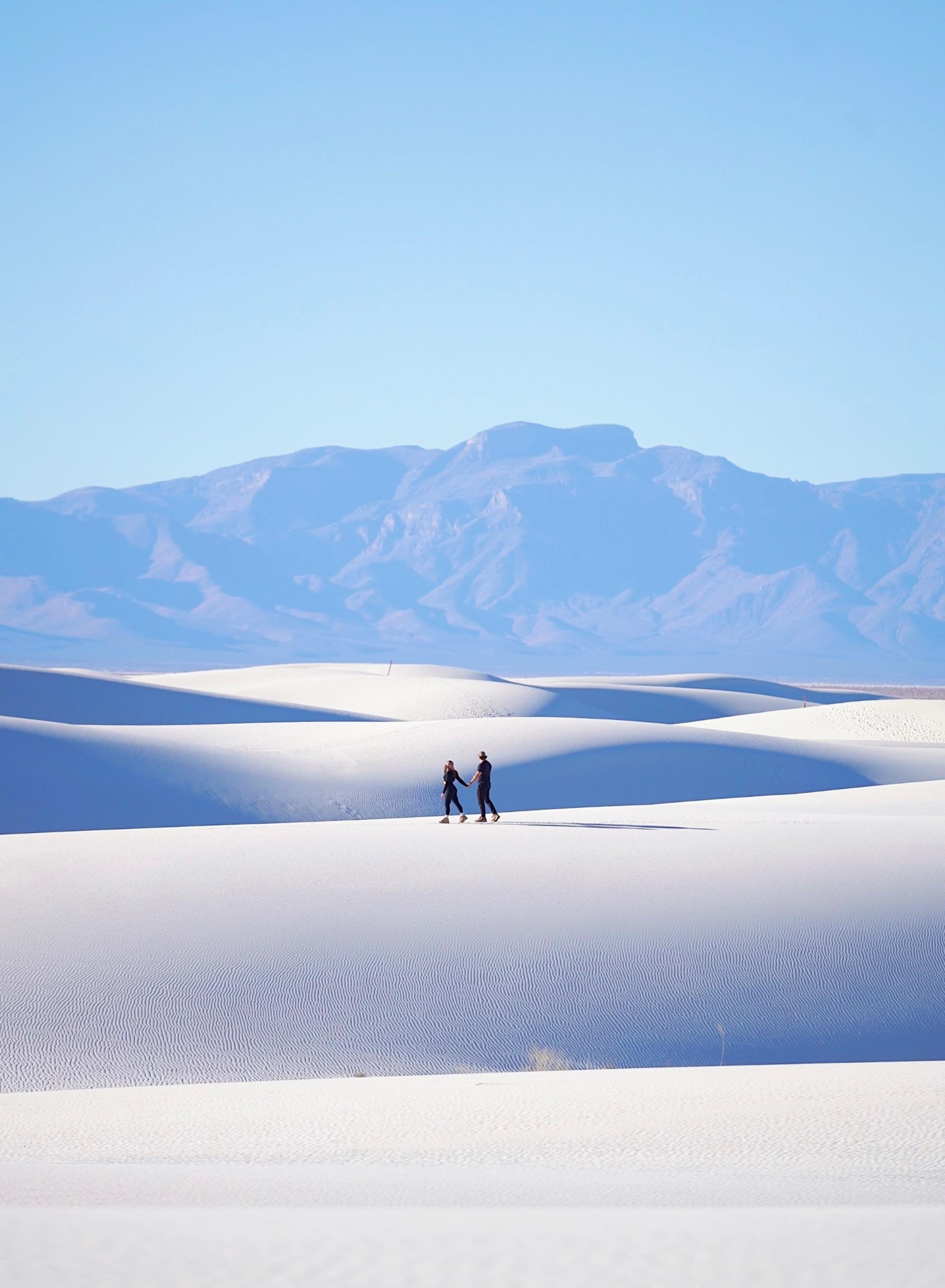 Two people hold hands while walking across vast white gypsum sand dunes, appearing small against the sweeping landscape. Blue mountain ranges stretch across the horizon under a clear sky, likely White Sands National Park, New Mexico.