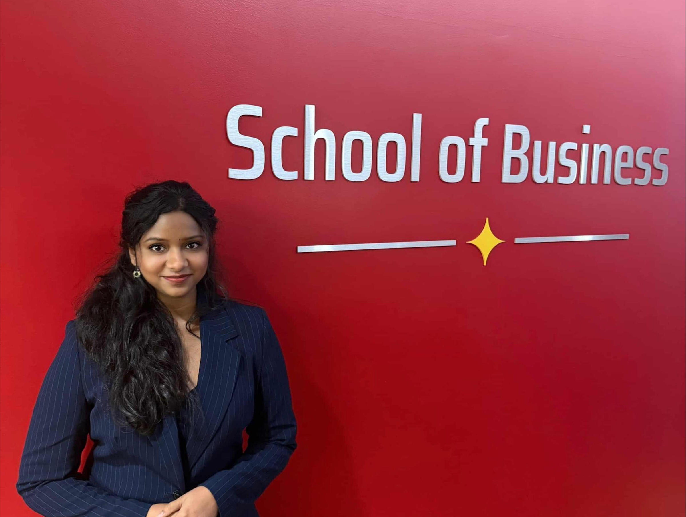 A woman with long dark hair in a navy pinstripe blazer poses in front of a red wall displaying "School of Business" signage with a decorative yellow star element. Professional portrait in what appears to be a university or business school setting.