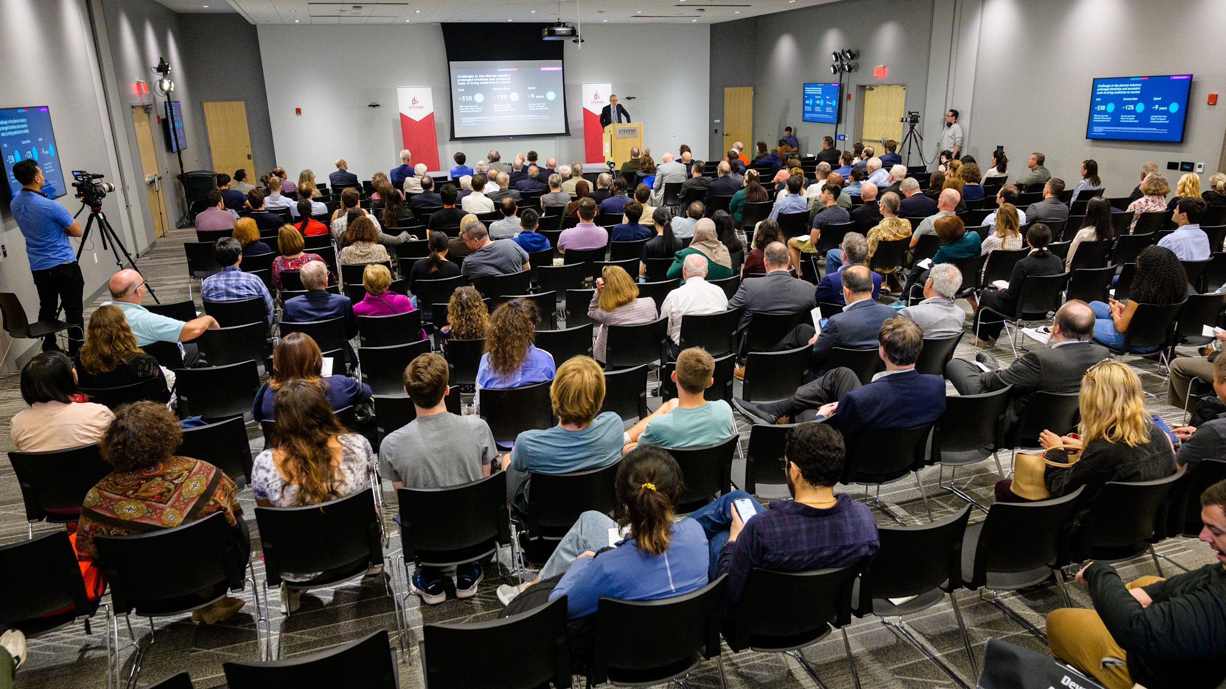 A full shot of a seated audience listening to a speaker give a presentation.
