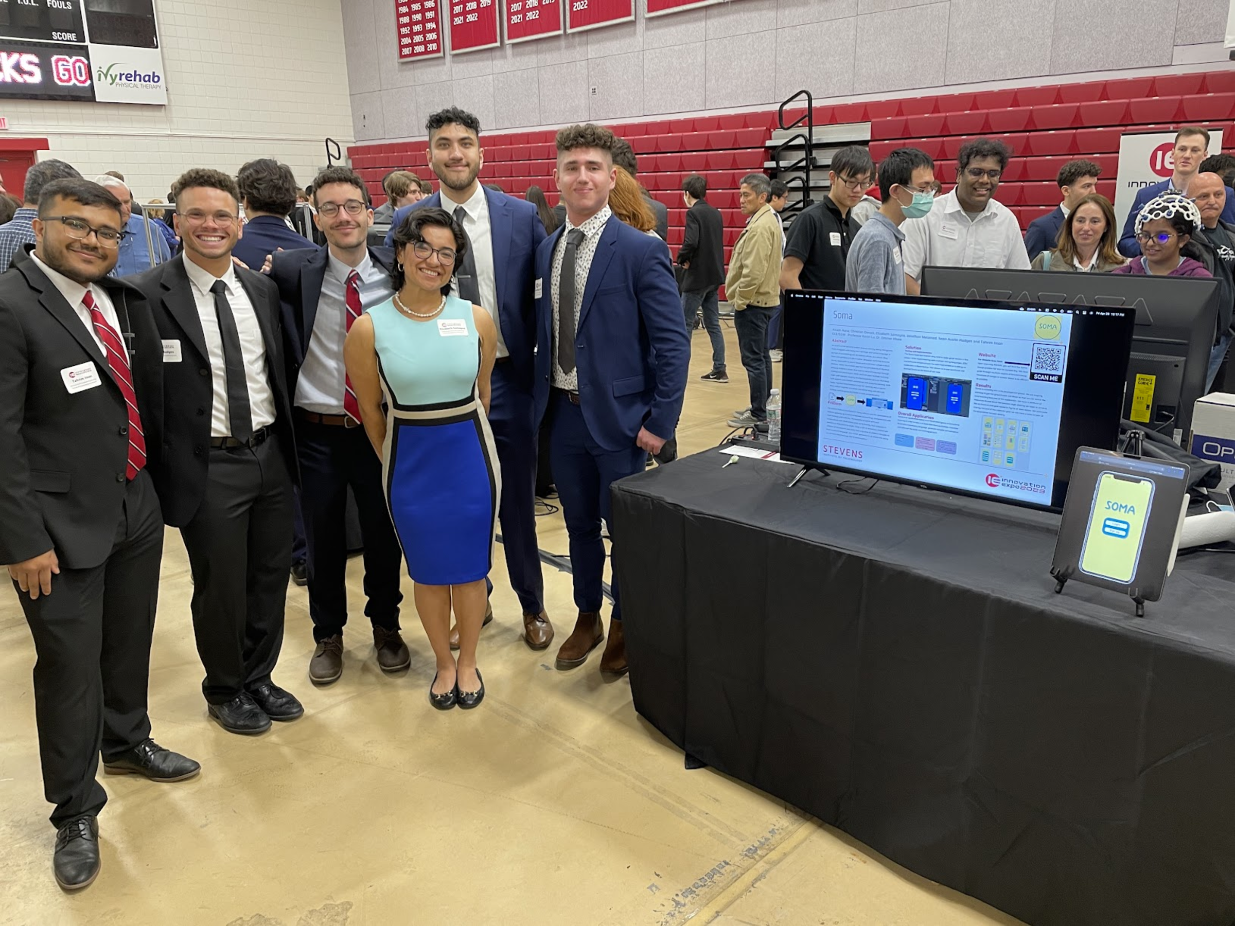 Soma team members stand next to a long table, which contains a computer screen showing their project poster presentation and a physical mock-up representing their Soma app. Other attendees of the 2023 Innovation Expo appear in the background.