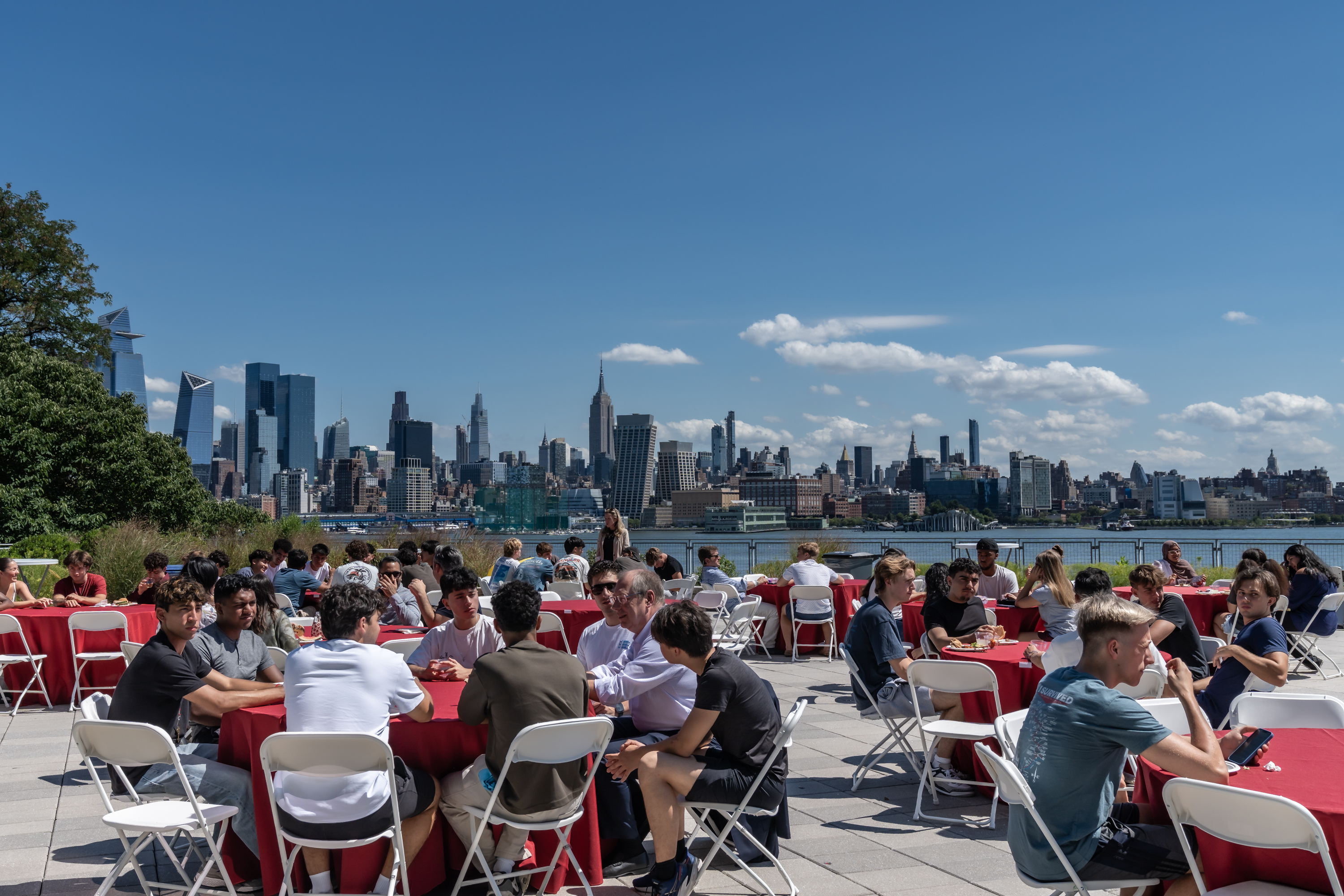 Outdoor dining event with red tablecloths and white folding chairs on waterfront terrace, Manhattan skyline visible across river on sunny day.
