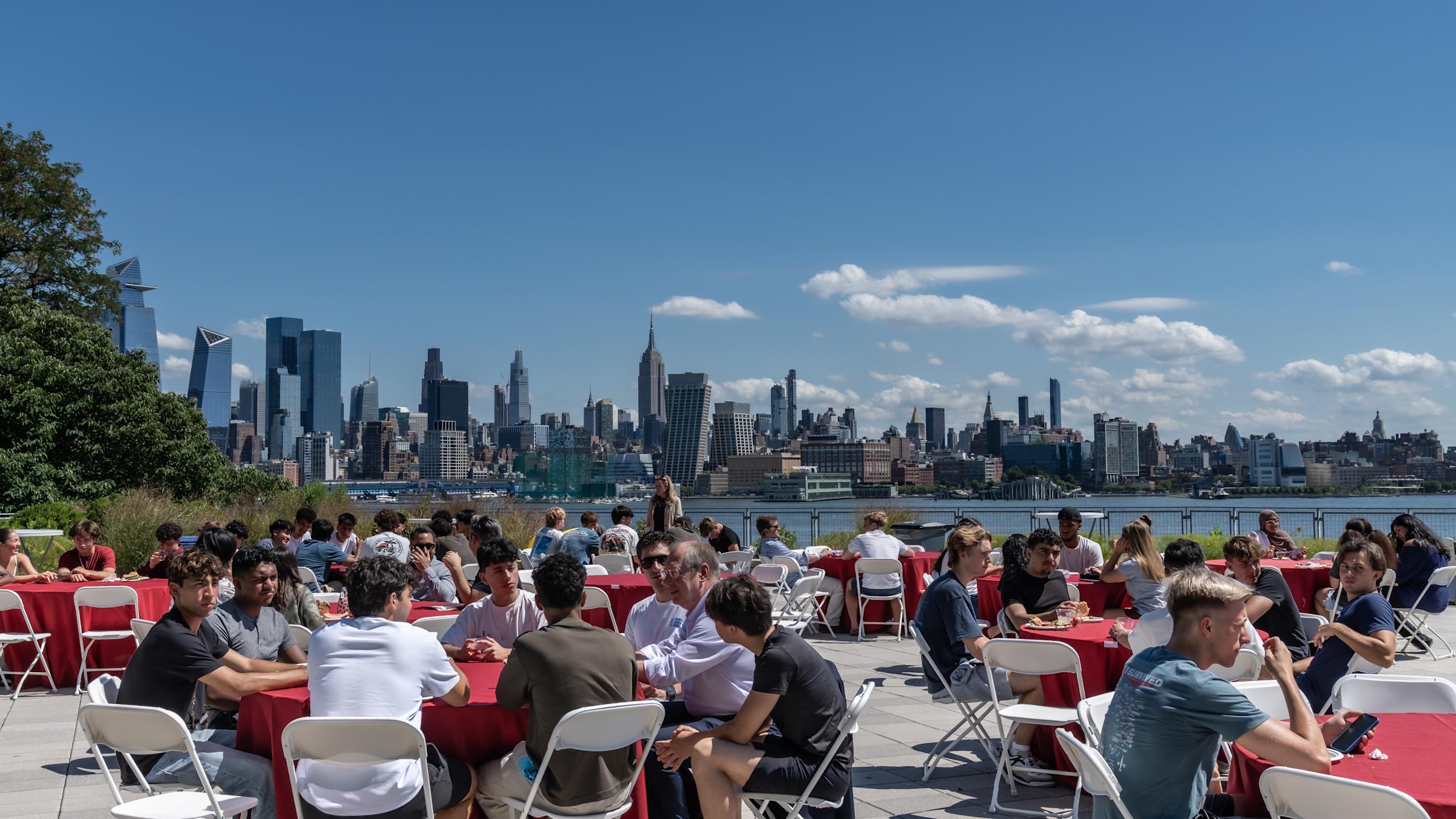 Outdoor dining event with red tablecloths and white folding chairs on waterfront terrace, Manhattan skyline visible across river on sunny day.