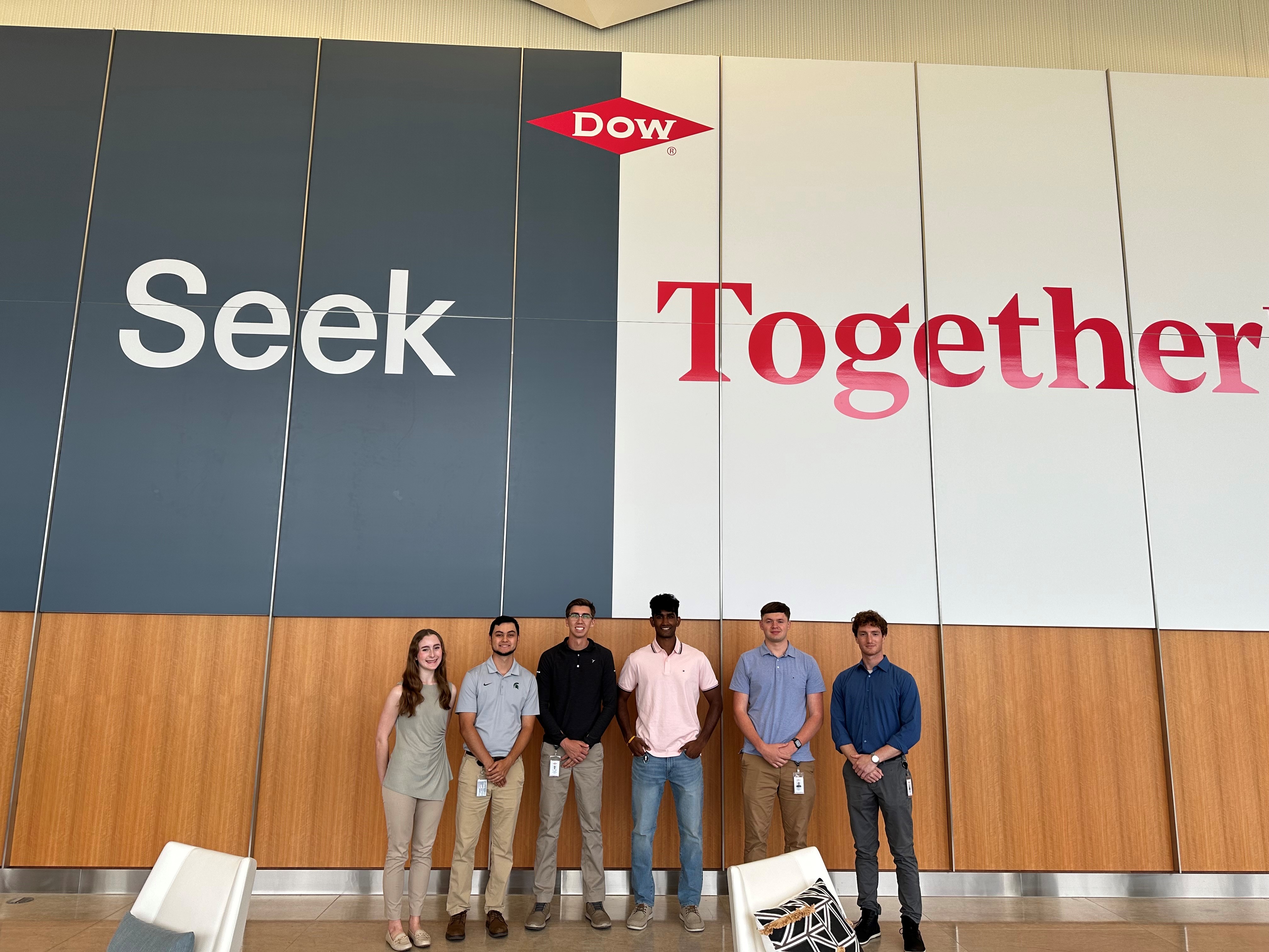 Group of interns stand in front of a large Dow sign that read, "Seek Together."