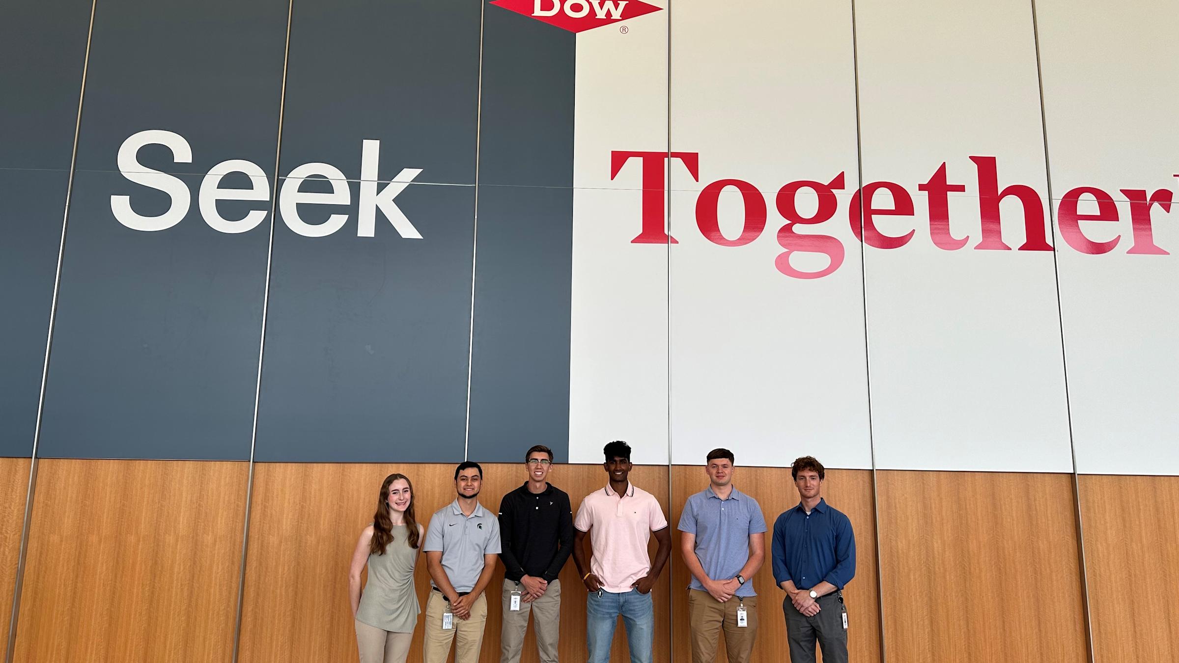 Group of interns stand in front of a large Dow sign that read, "Seek Together."