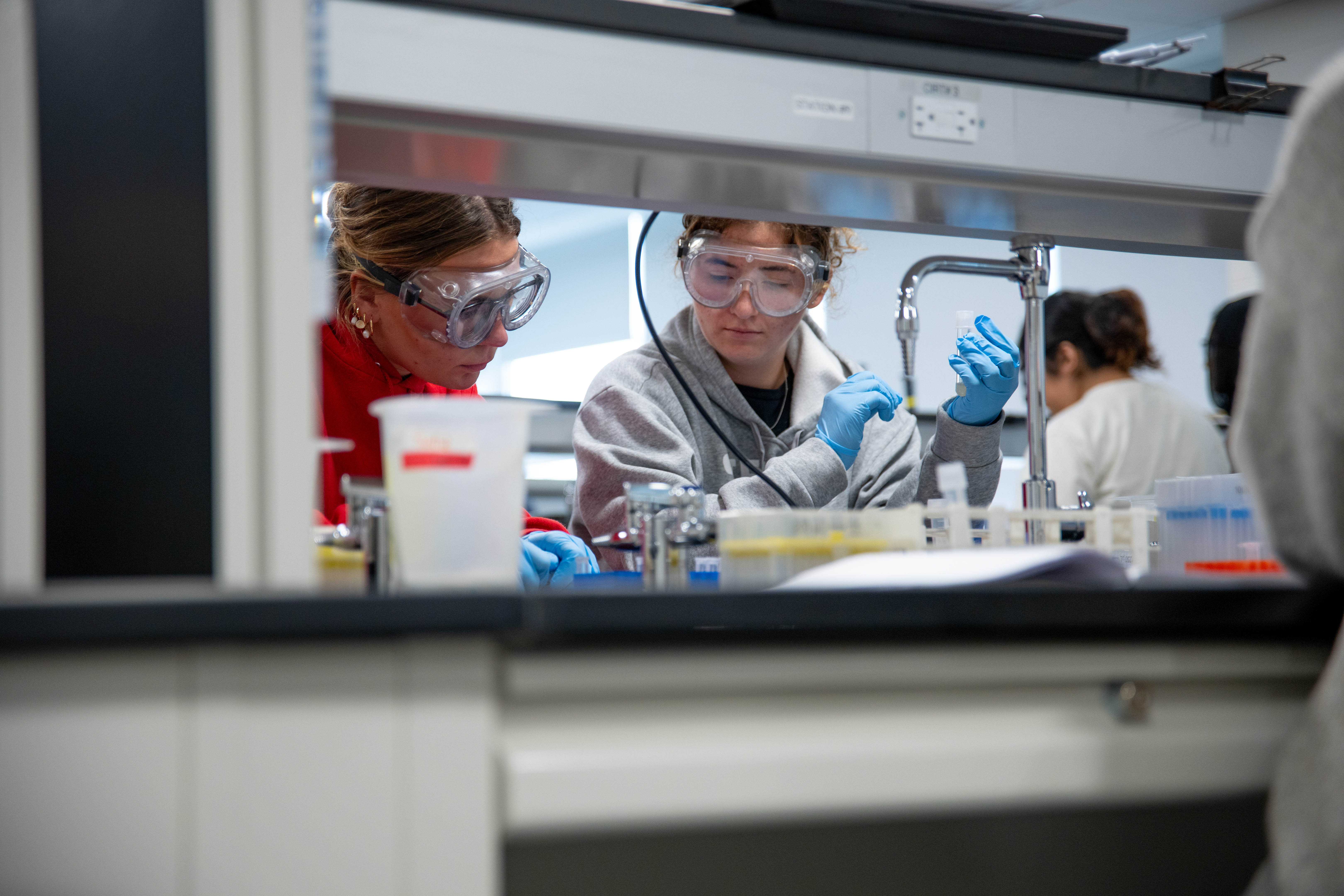 Two female students in protective googles, lab coats and gloves work in a lab