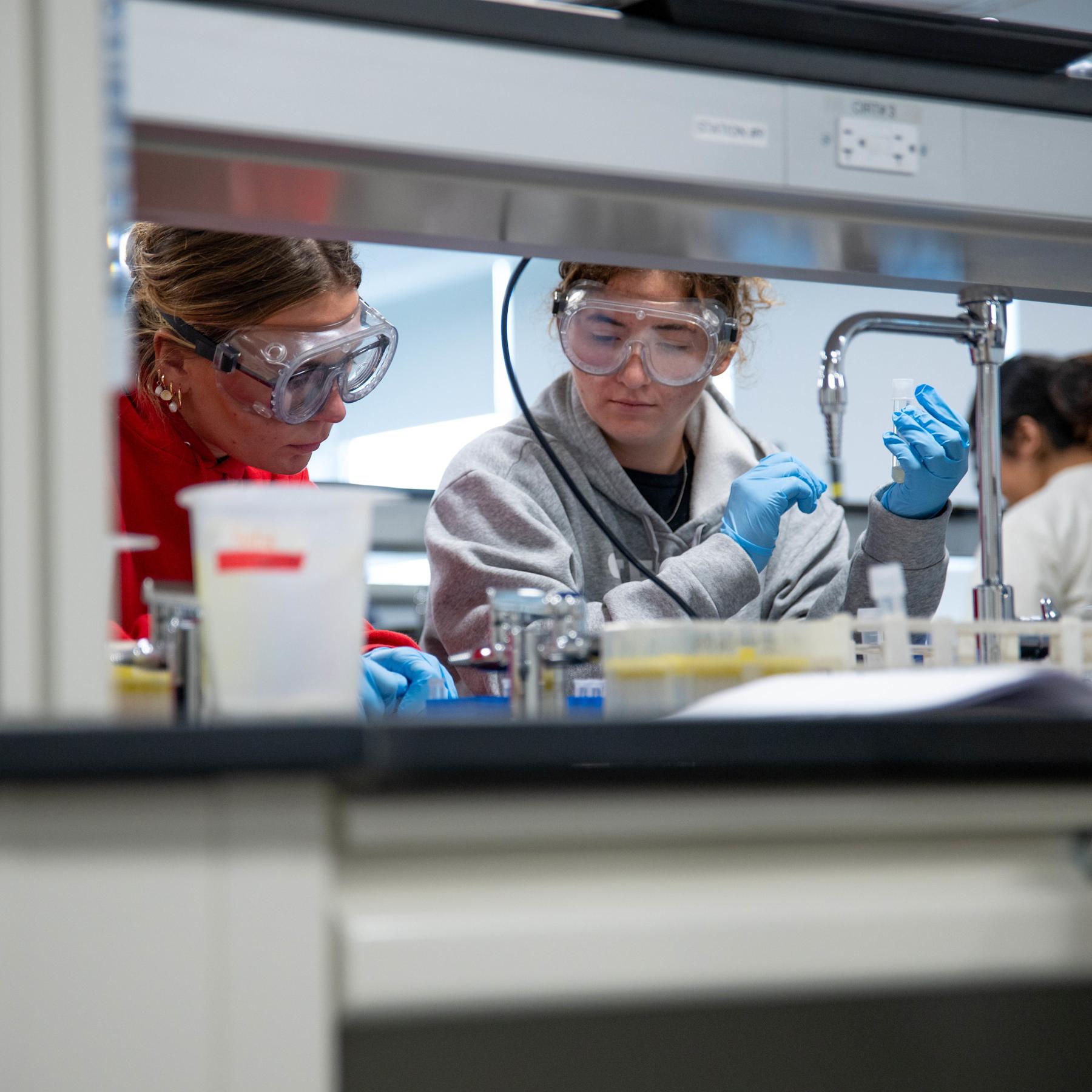 Two female students in protective googles, lab coats and gloves work in a lab