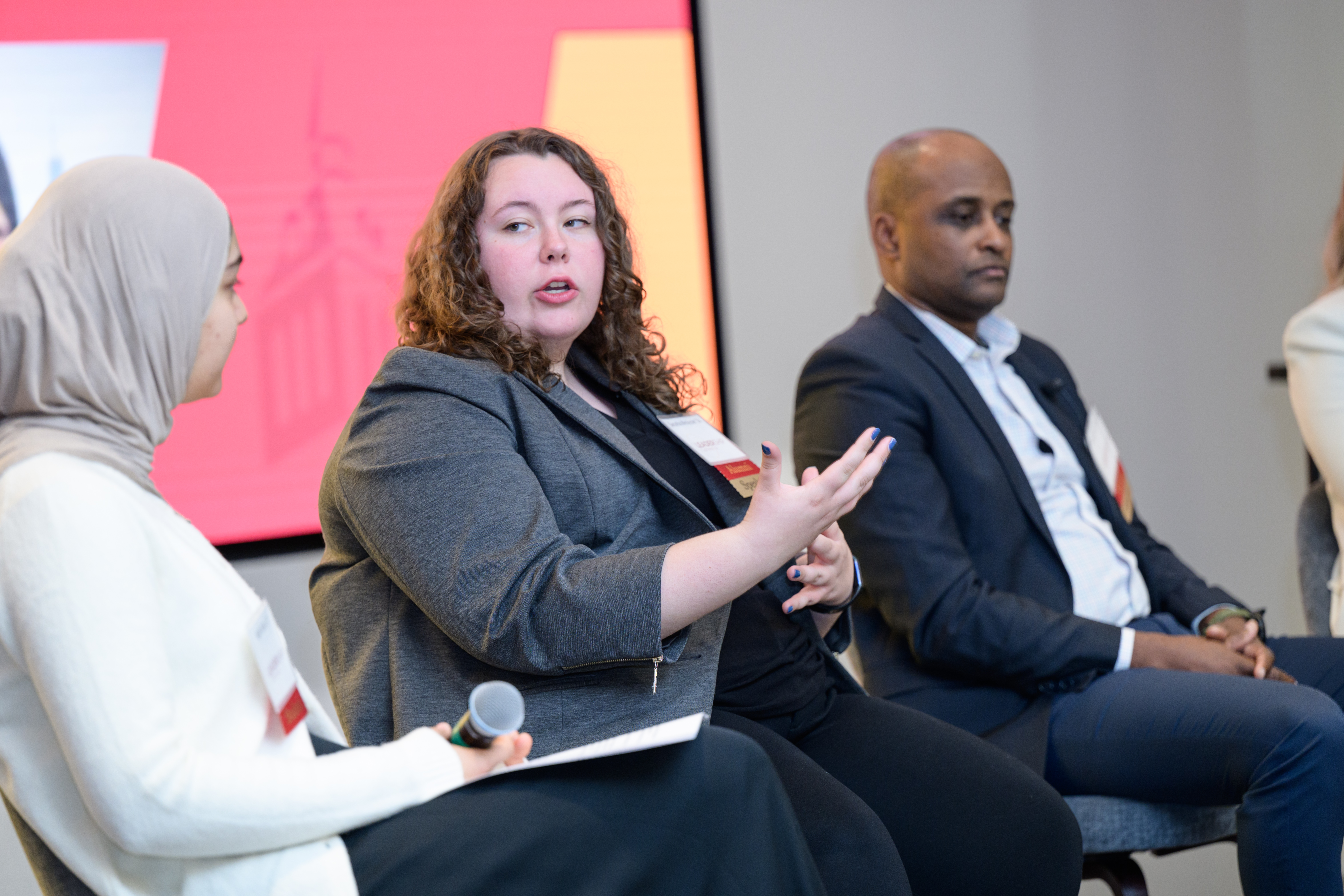 Three individuals participate in a panel discussion while seated on stage. The person in the center, wearing a gray blazer, gestures while speaking, while another panelist on the left holds a microphone and a notepad. 