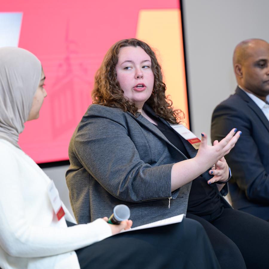 Three individuals participate in a panel discussion while seated on stage. The person in the center, wearing a gray blazer, gestures while speaking, while another panelist on the left holds a microphone and a notepad.