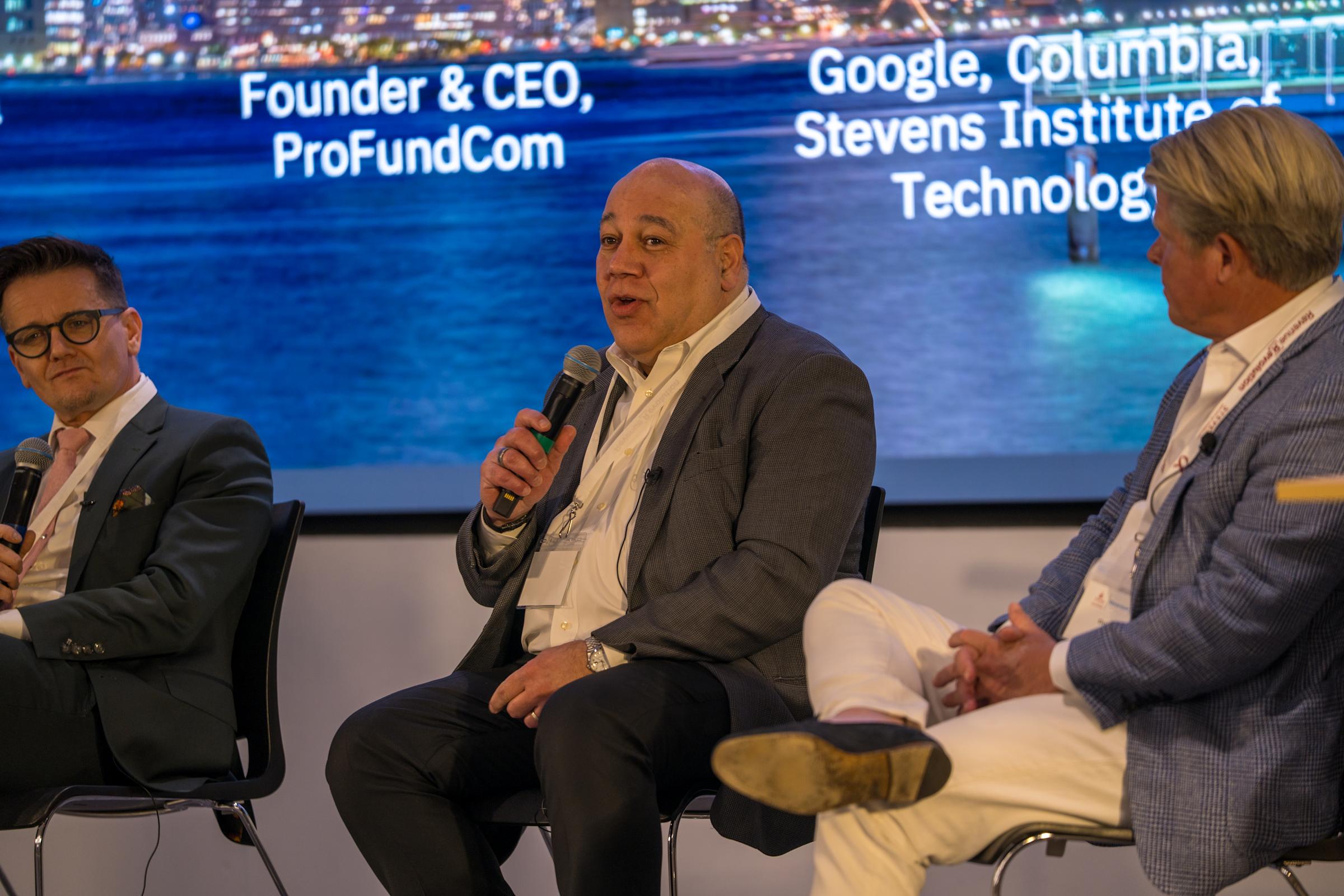 Three executives in business attire sit on stage for a panel discussion, with the center speaker - Founder & CEO of ProFundCom - holding a microphone. .