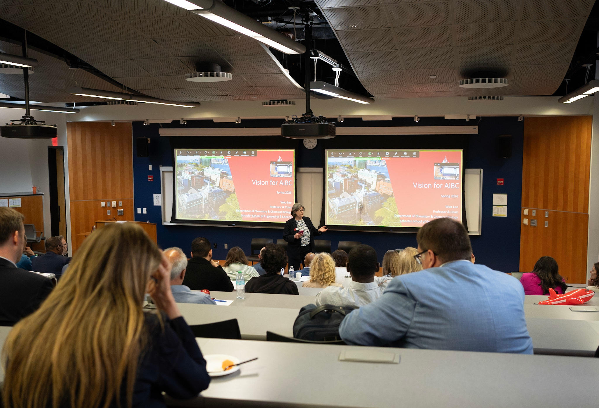 Patricia Muisener delivering a lecture in front of two screens in an elevated lecture hall with attendees.