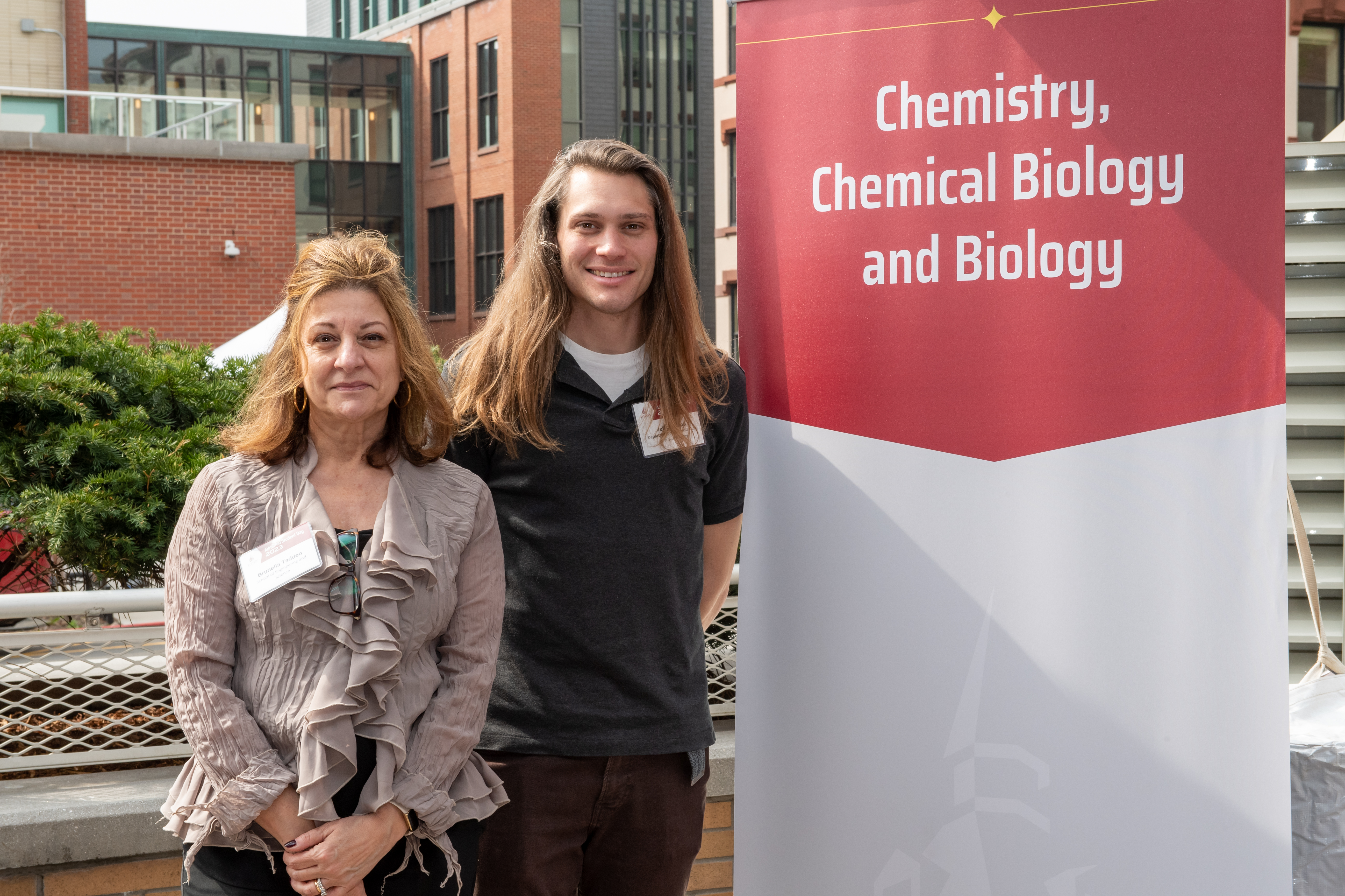 Professor Brunella Taddeo and Lecturer Jeffrey Raab posing for a picture on campus next to a banner that says Chemistry, Chemical Biology and Biology