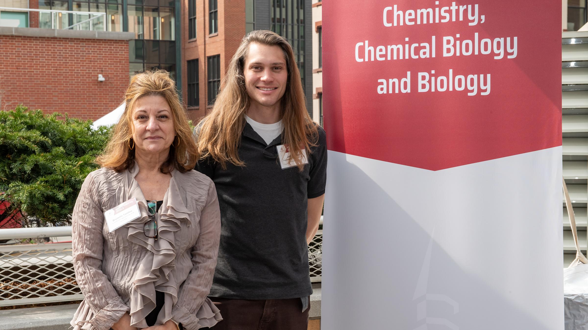 Professor Brunella Taddeo and Lecturer Jeffrey Raab posing for a picture on campus next to a banner that says Chemistry, Chemical Biology and Biology