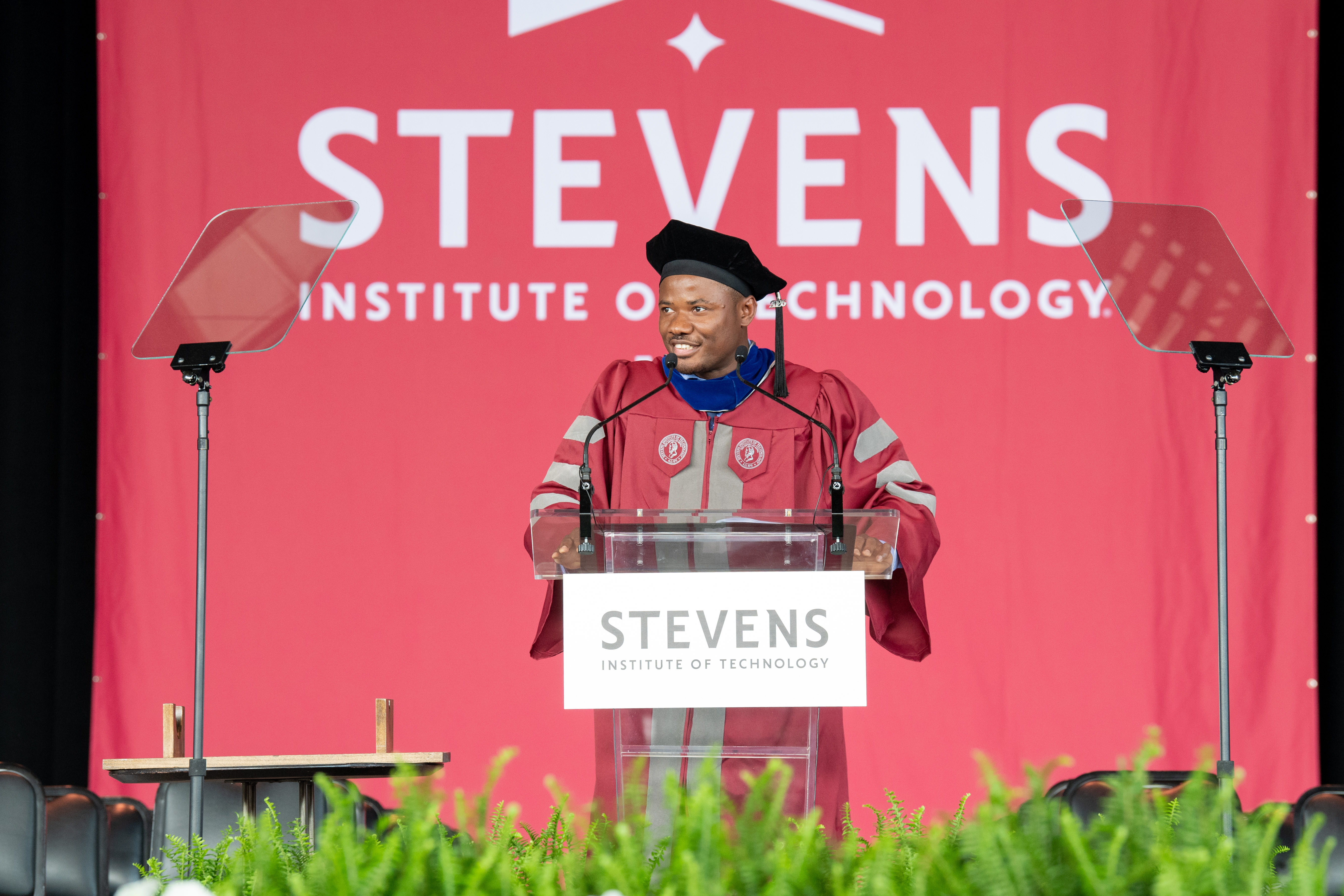 Christopher Mbonu standing at a clear podium on a stage for graduation, in traditional graduation cap and gown.