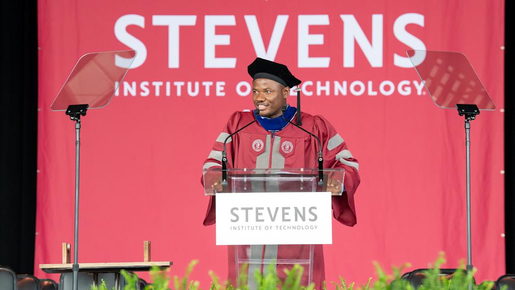 Christopher Mbonu standing at a clear podium on a stage for graduation, in traditional graduation cap and gown.