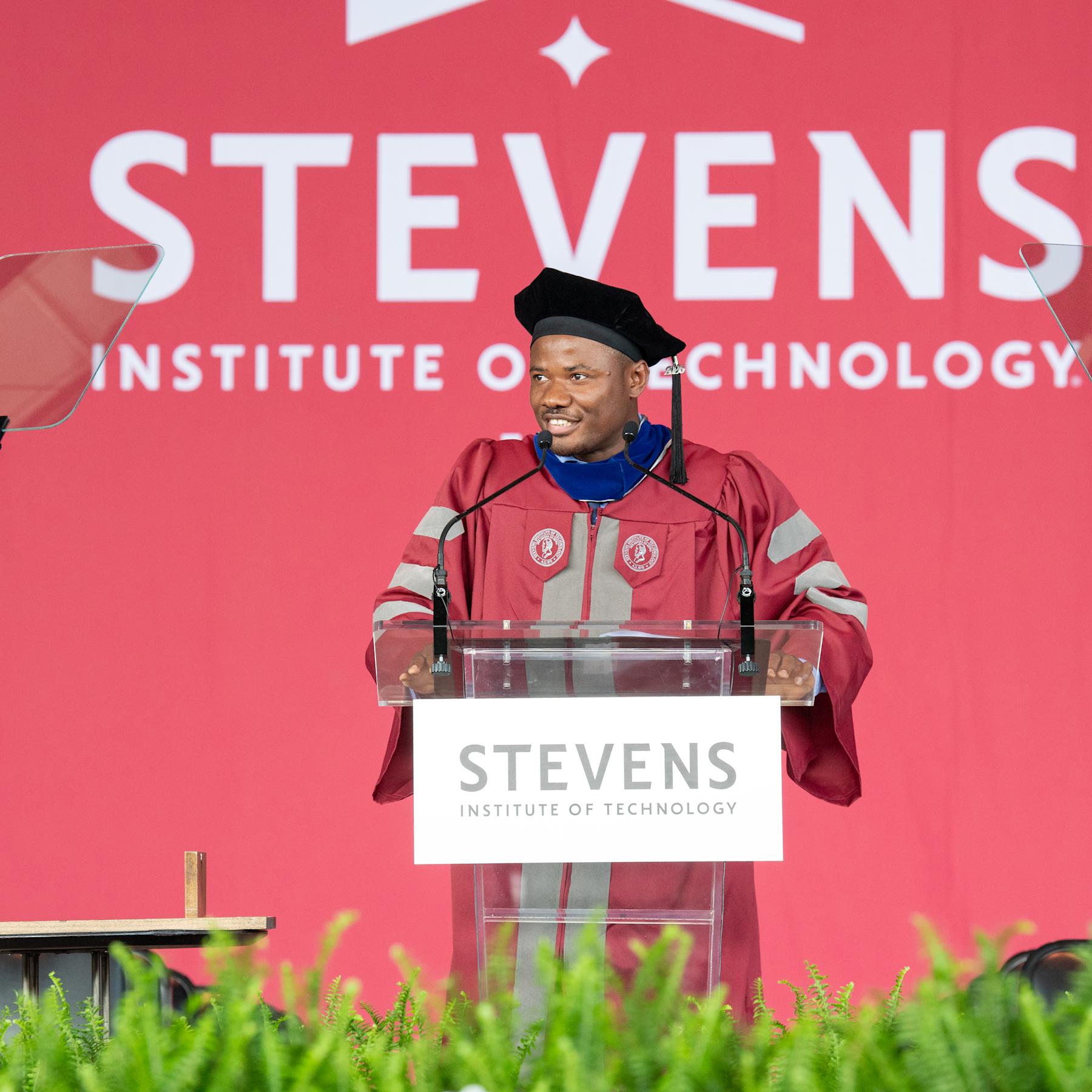 Christopher Mbonu standing at a clear podium on a stage for graduation, in traditional graduation cap and gown.