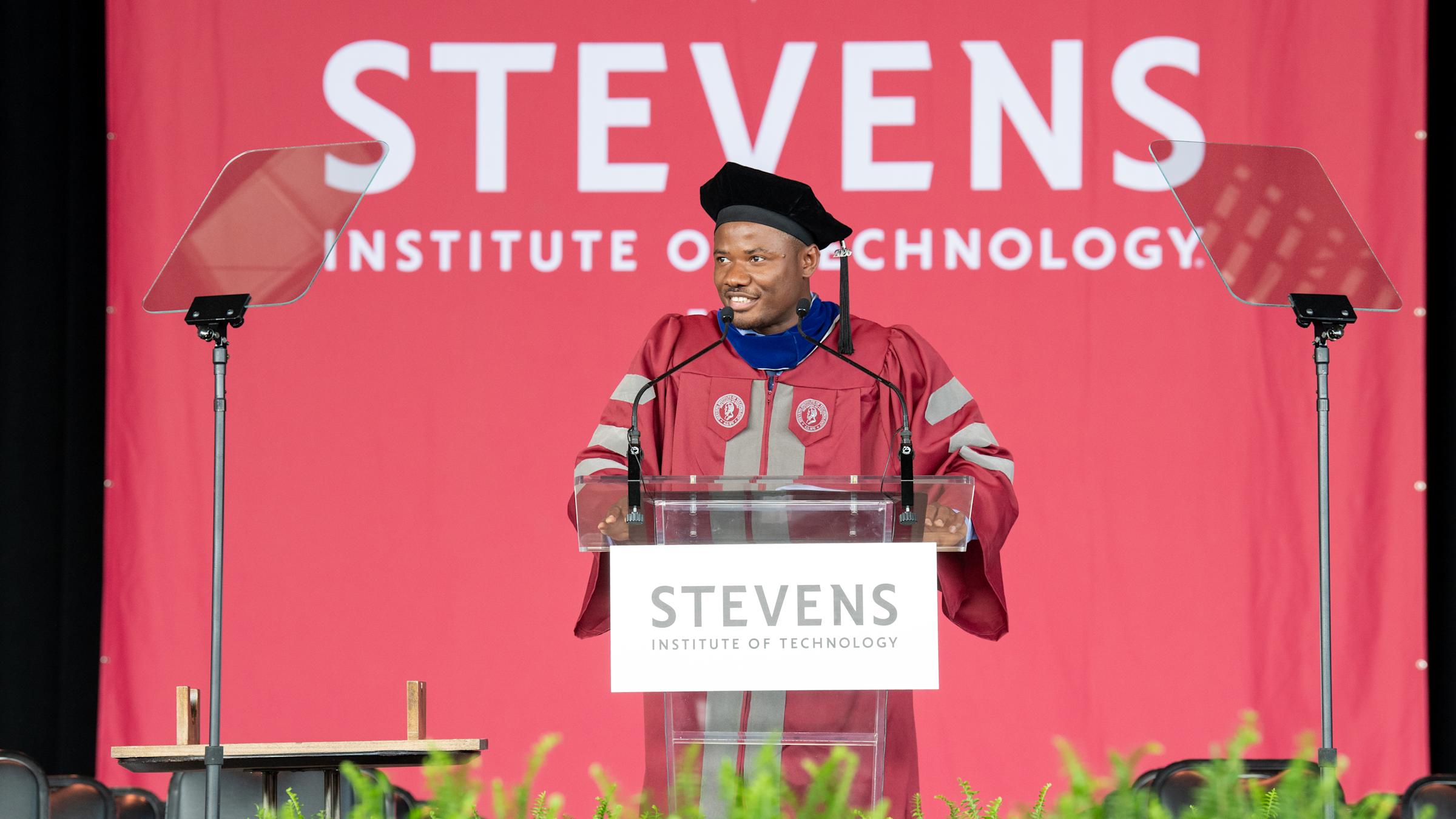 Christopher Mbonu standing at a clear podium on a stage for graduation, in traditional graduation cap and gown.