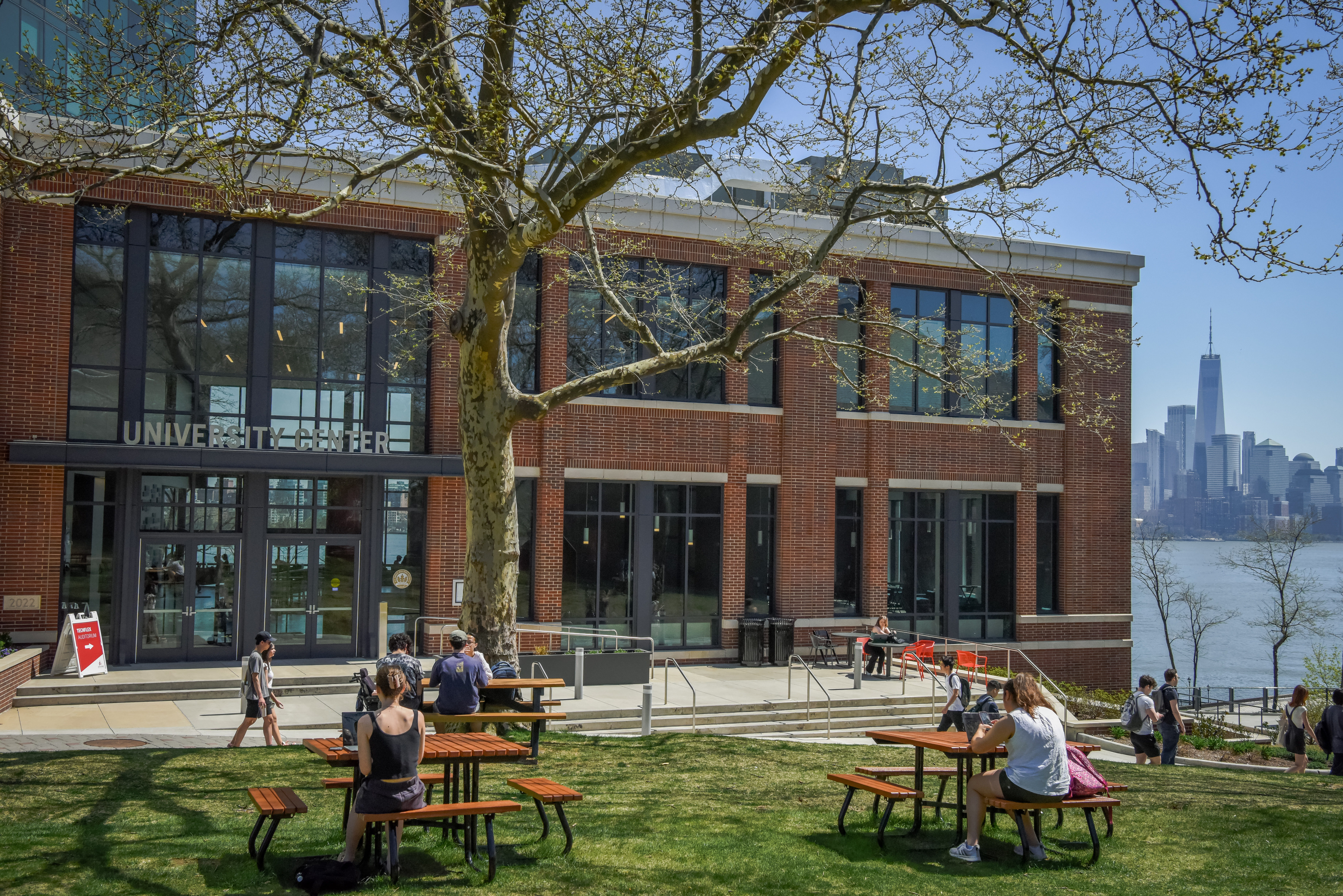 Students studying on the lawn and at picnic tables in front of the University Center with One World Trade in the background.