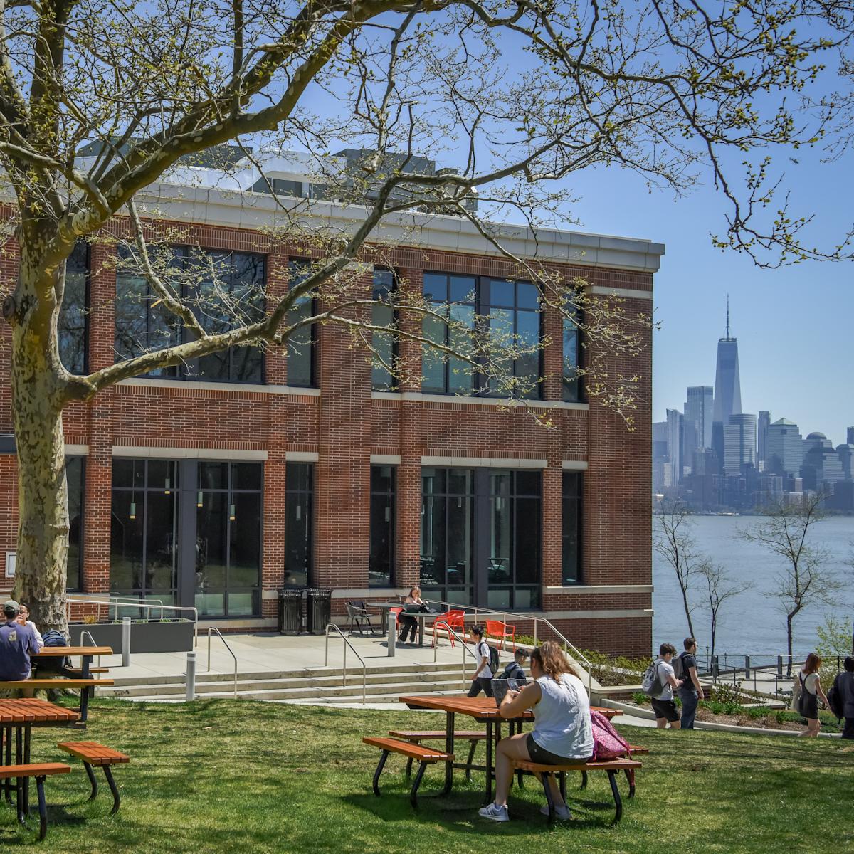 Students studying on the lawn and at picnic tables in front of the University Center with One World Trade in the background.