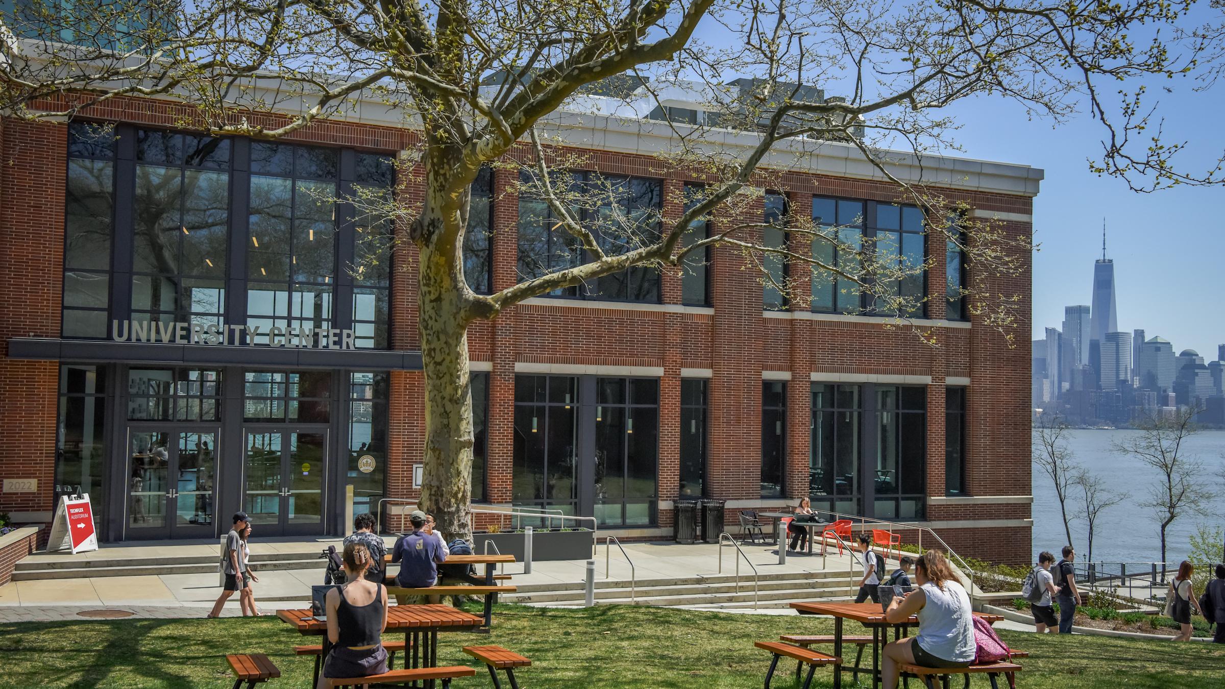 Students studying on the lawn and at picnic tables in front of the University Center with One World Trade in the background.
