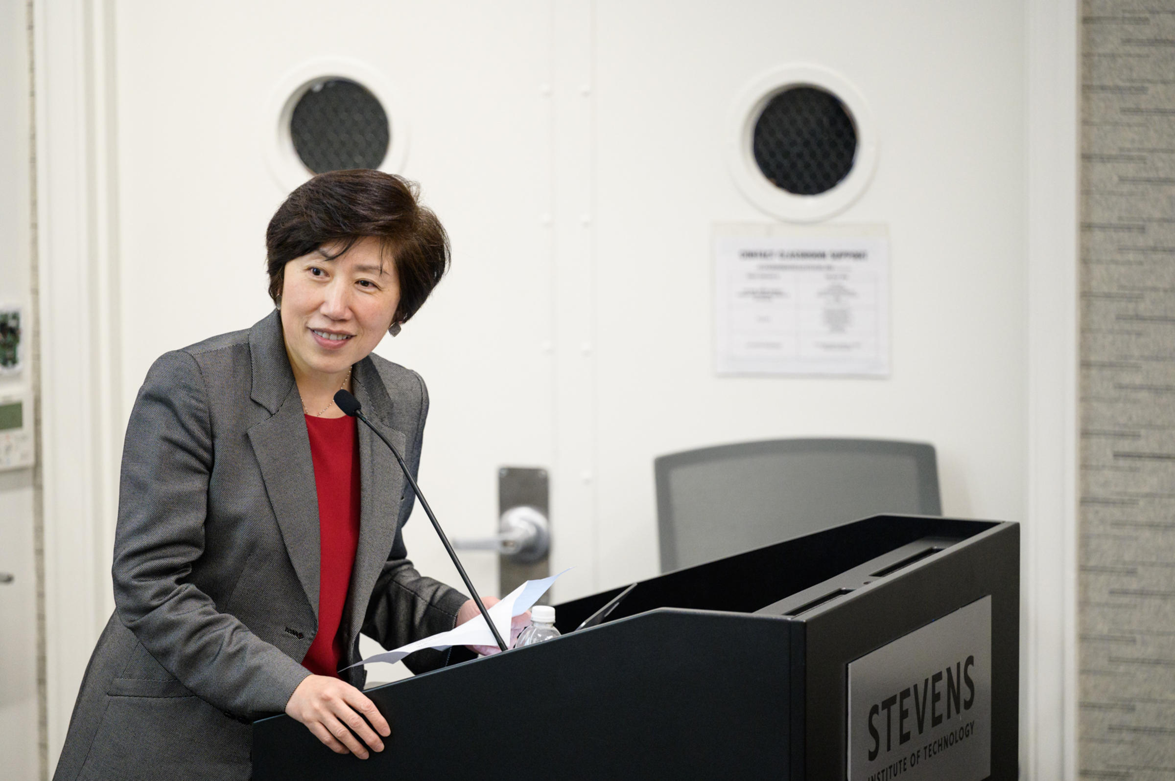 Jean Zu stands behind a podium over a microphone looking over her shoulder and speaking
