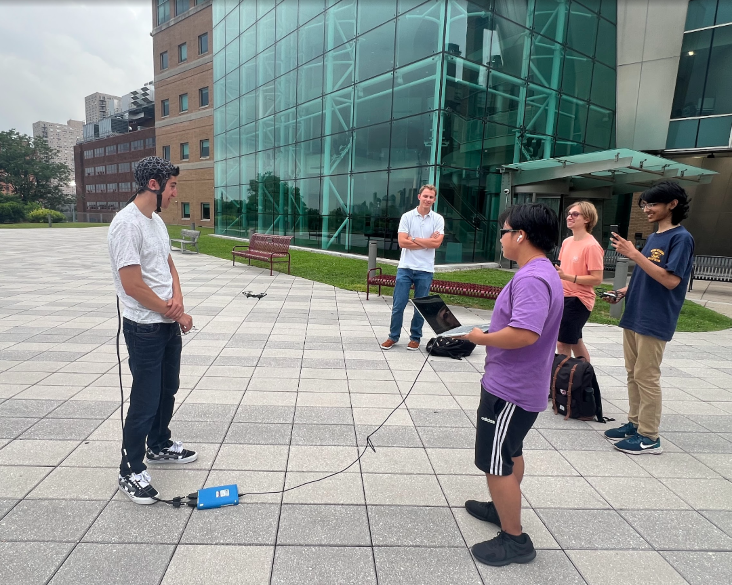 Students standing in front of a building with drone equipment that use wearable head devices that read brain waves.