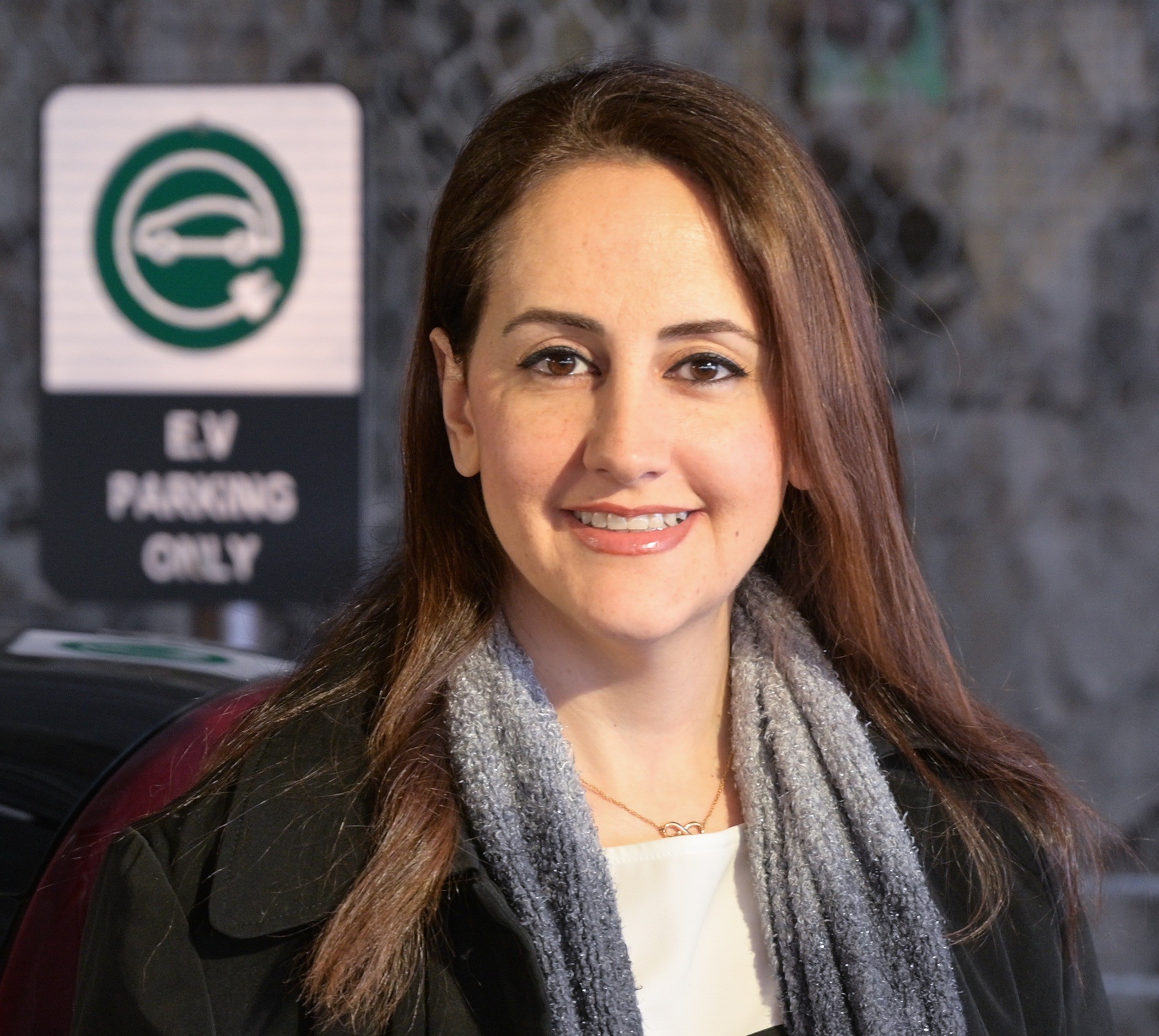 A headshot of Yeganeh Hayeri in front of an electric vehicle sign