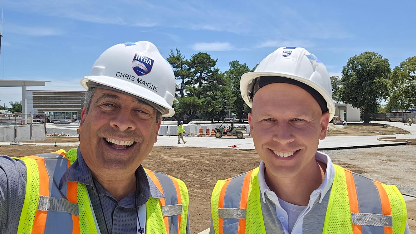 Christopher Maione ’82 and Steven Koch ’12 wear hard hats and safety vests while smiling outside at the Belmont Park construction site.
