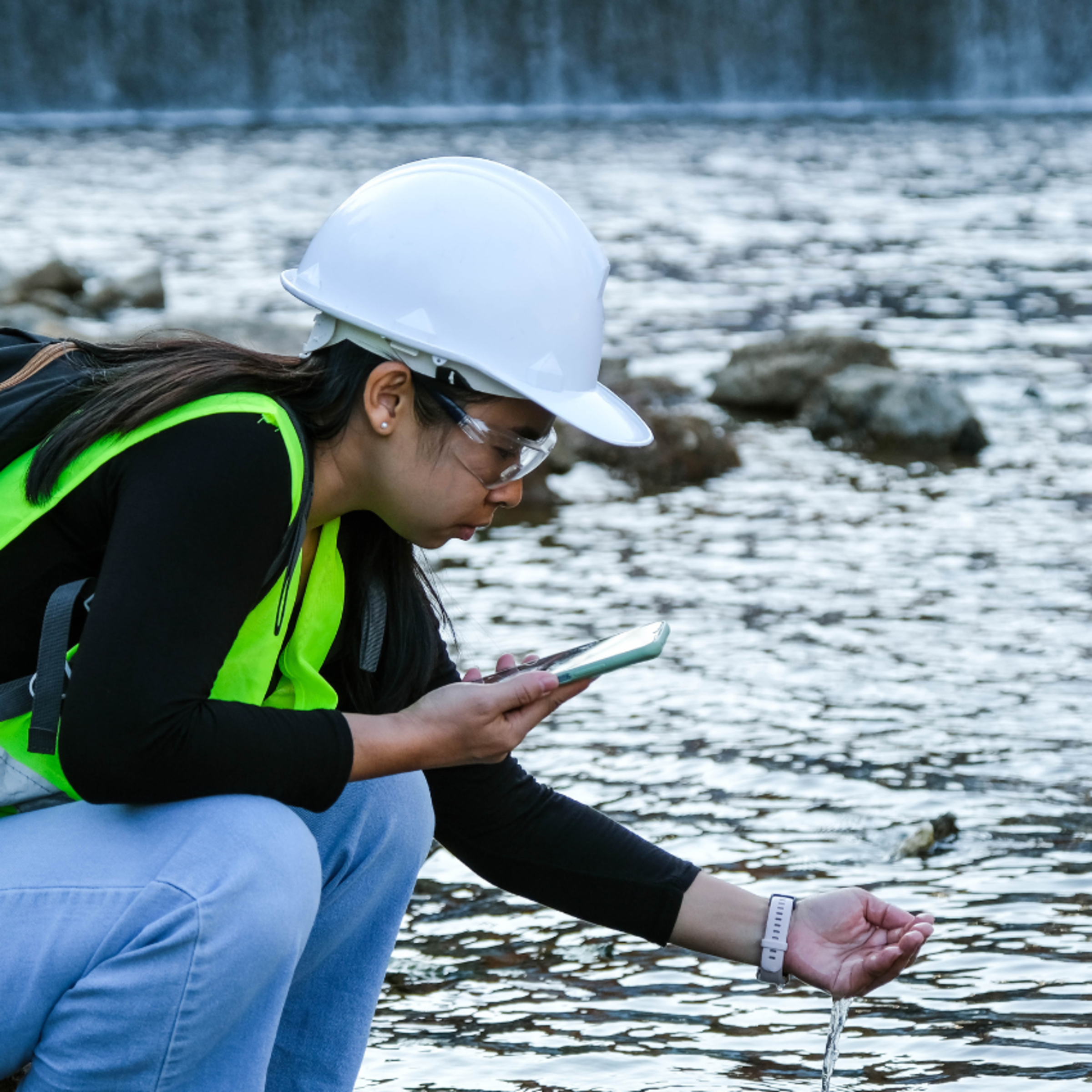 An environmental engineering student in a hard helmet and vest testing water samples with a phone.