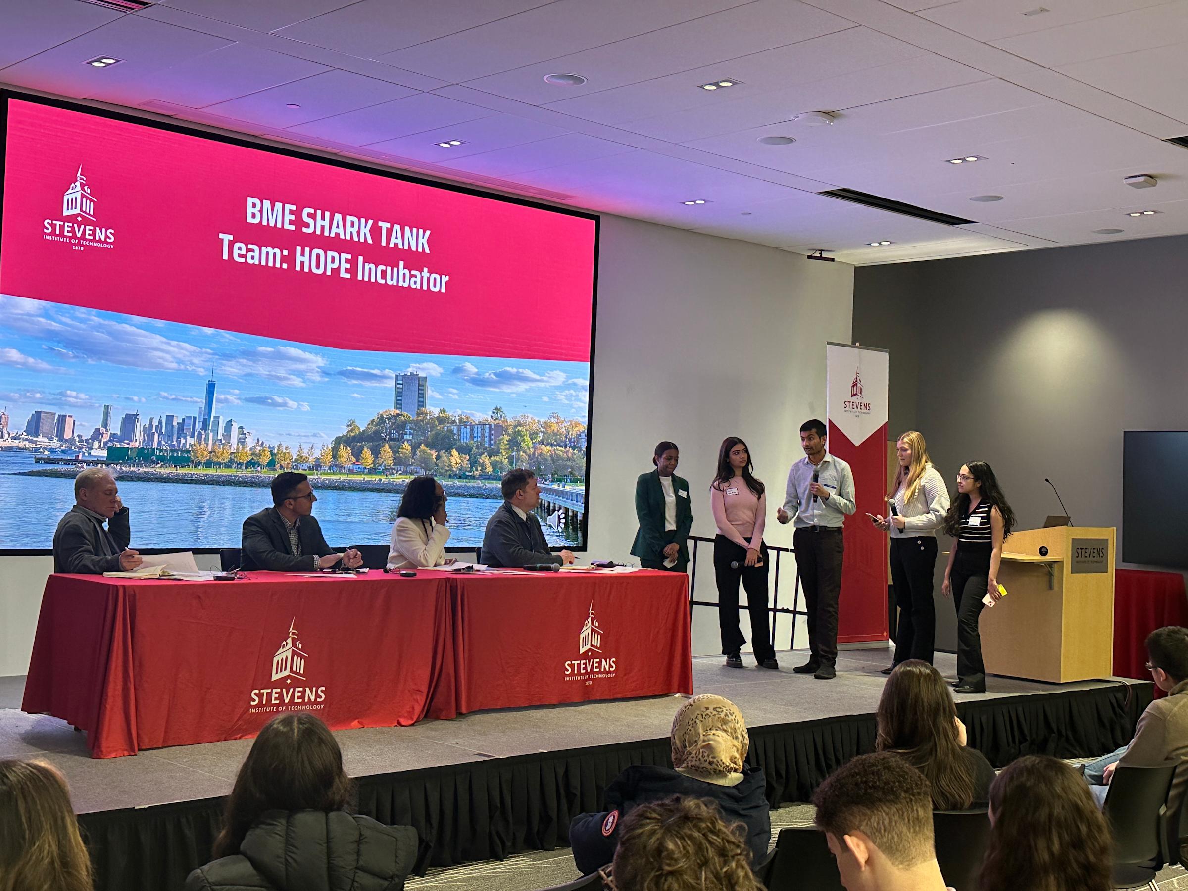A group of five biomedical engineering students standing on a presenting platform next to a panel of judges presenting their project.