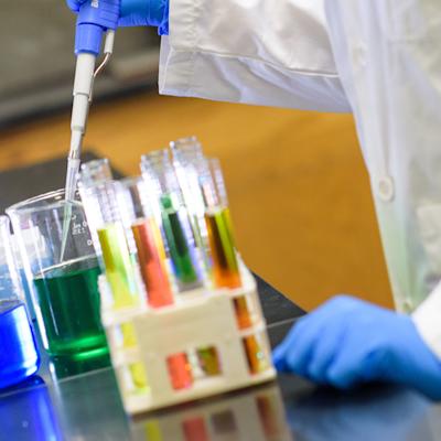 A researcher testing liquids in different sized beakers with liquids that are different colors wearing protective latex gloves and a lab coat.