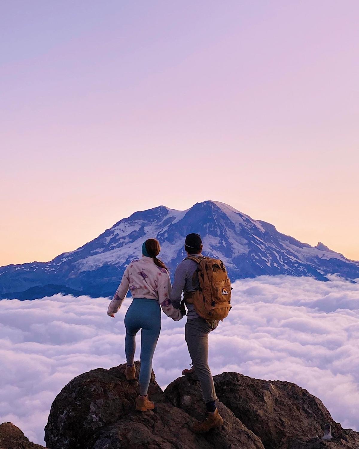 Two hikers hold hands on a rocky summit above a sea of clouds, gazing at a snow-capped mountain peak bathed in soft pink and golden sunset light. One wears a tan backpack and black cap; the other wears a tie-dye hoodie and teal leggings.