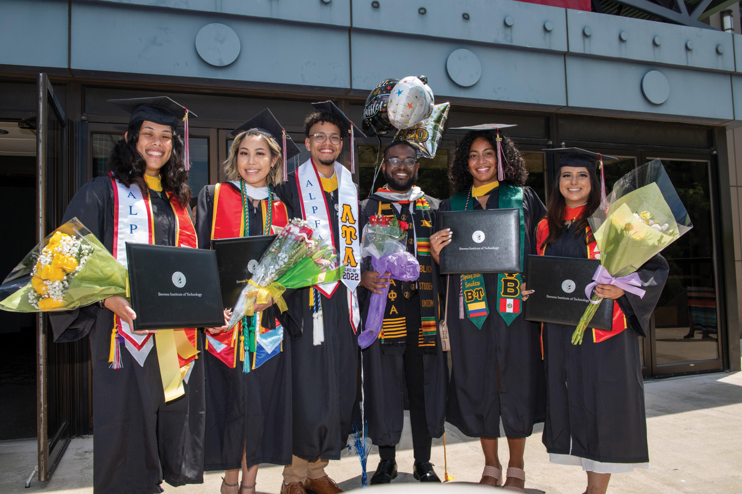 Graduating students in caps and gowns smile for the camera outside commencement venue.