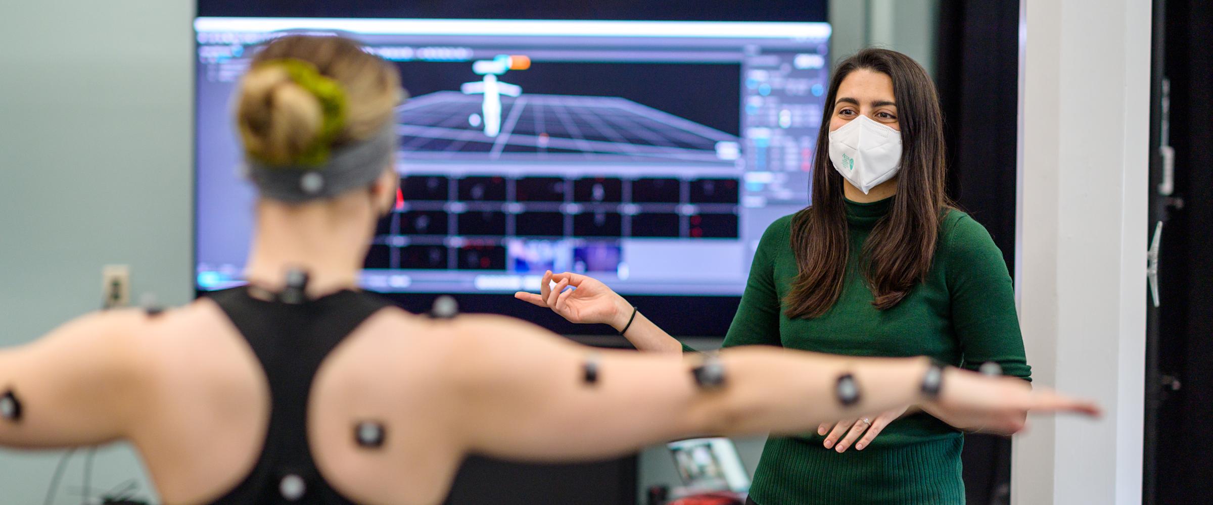 Model with motion capture nodes attached to arms raises arms with instructor in background