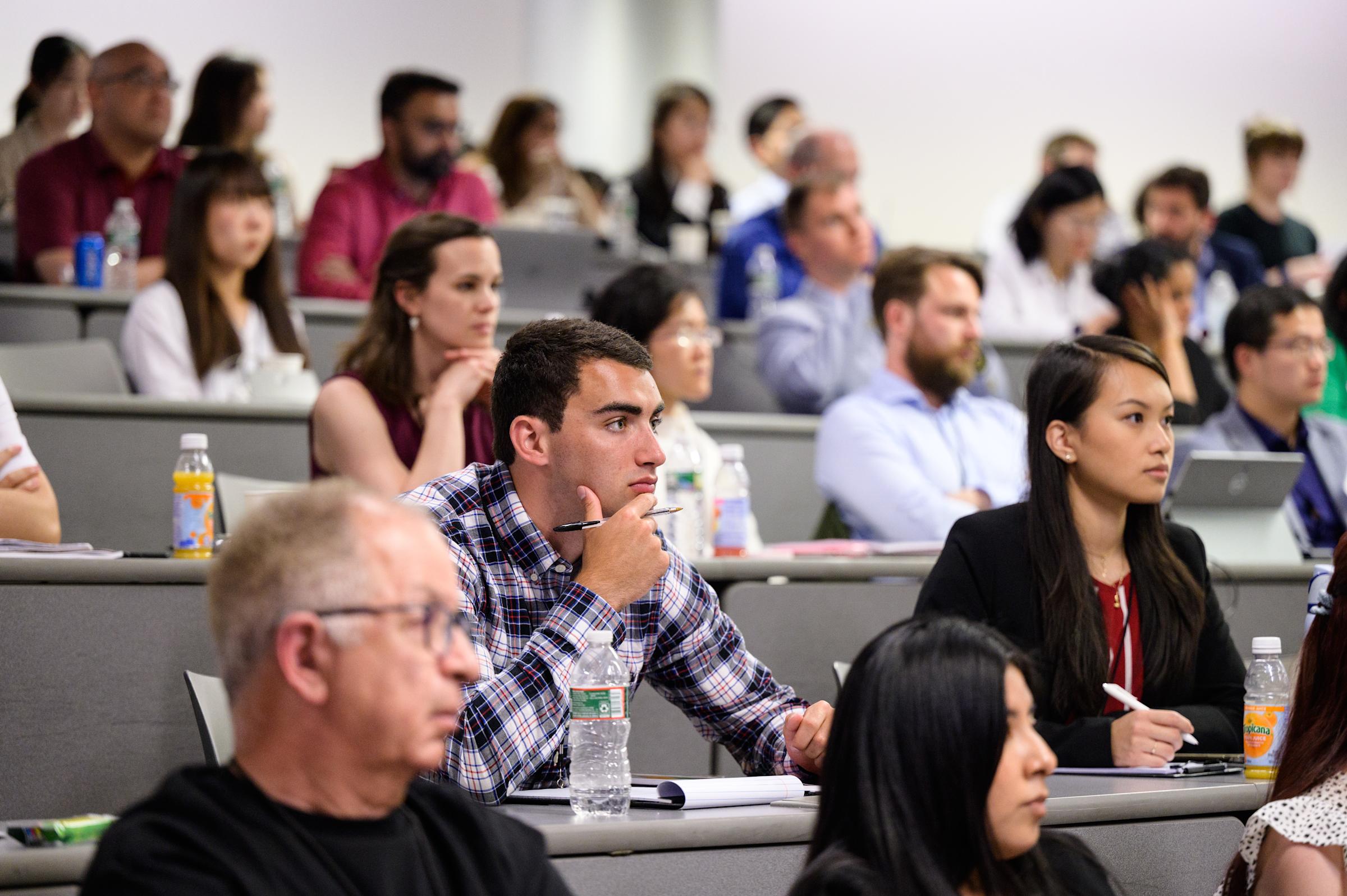 Professionally dressed people sit at tiered desks looking forward at (unseen) presenter
