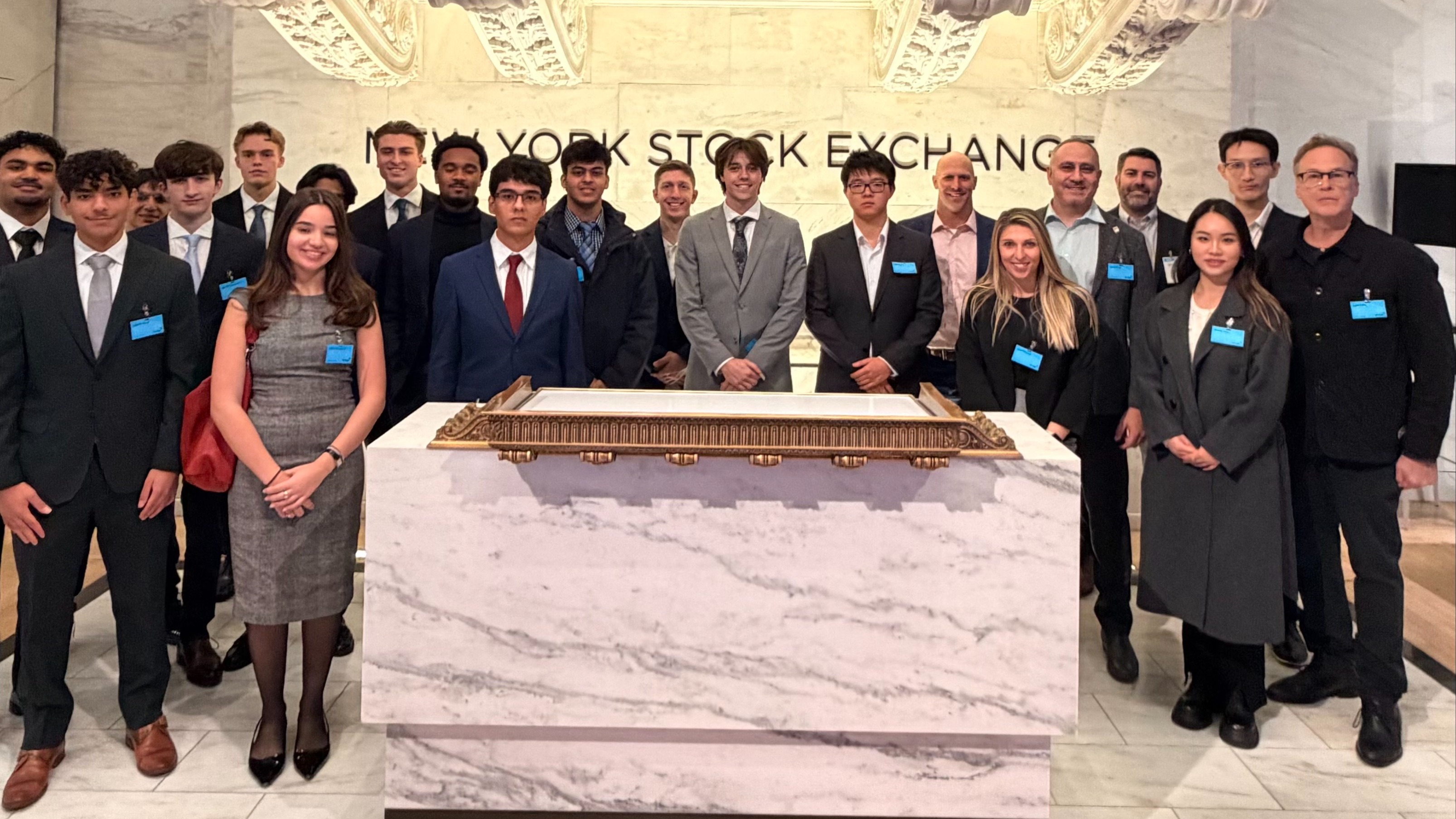 Large group of business students in formal attire pose at the New York Stock Exchange trading floor beneath the iconic brass bell and NYSE signage. The diverse group wears blue name badges and stands behind the marble trading desk.