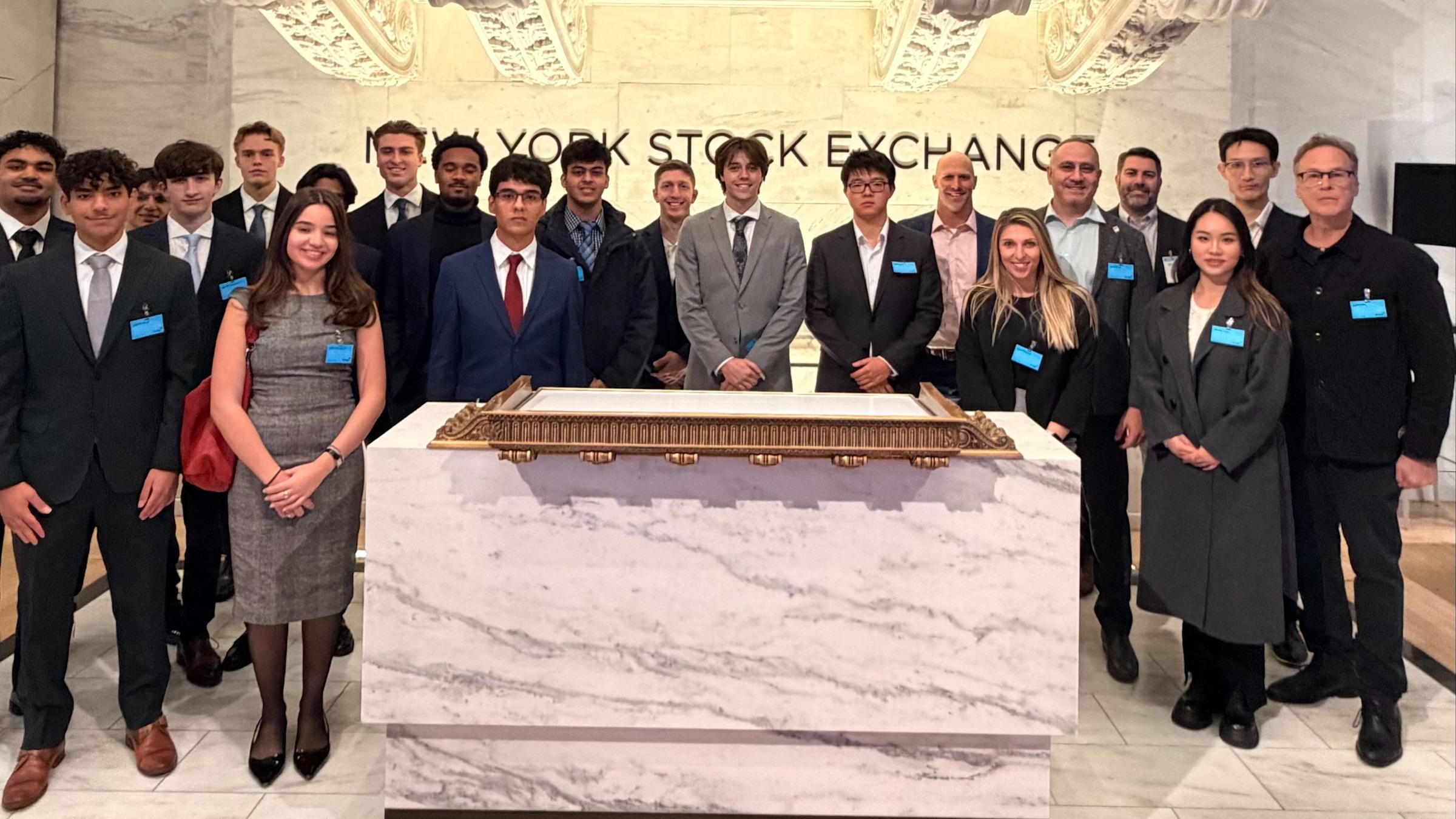 Large group of business students in formal attire pose at the New York Stock Exchange trading floor beneath the iconic brass bell and NYSE signage. The diverse group wears blue name badges and stands behind the marble trading desk.