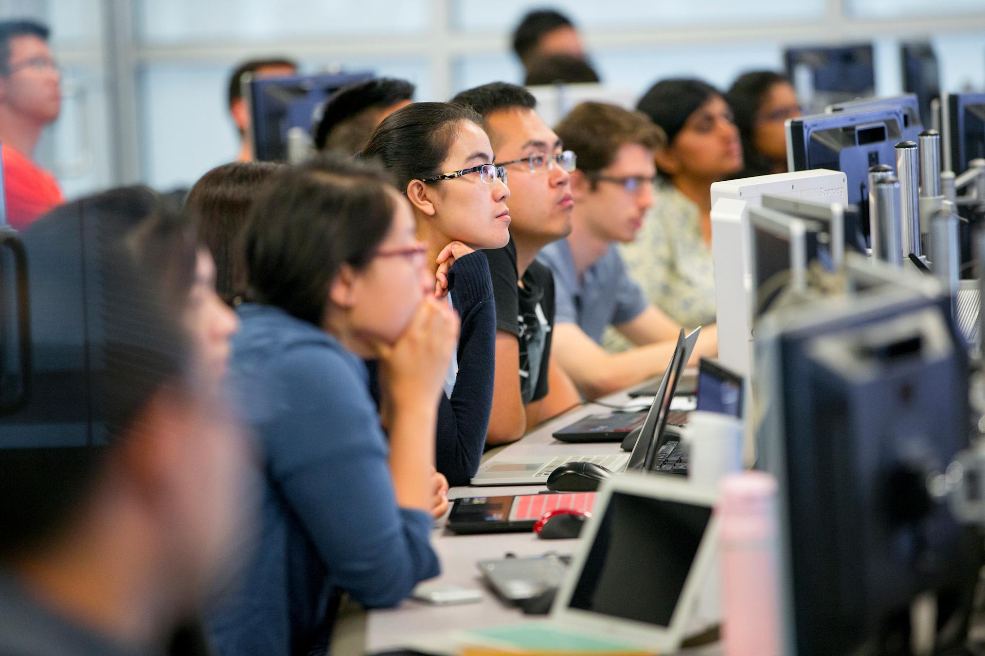 Students in front of computer monitors listen to instructor 