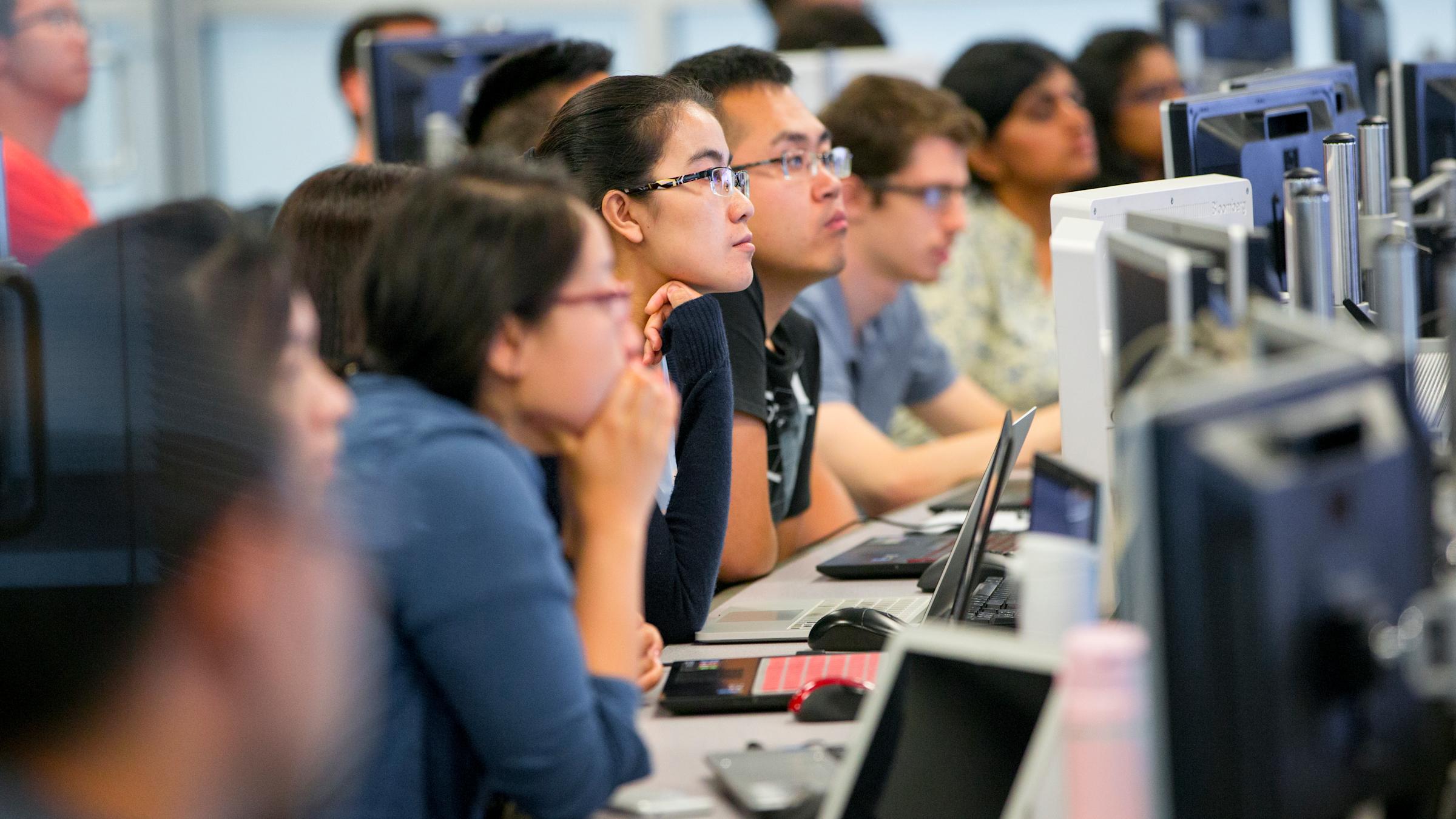 Students in front of computer monitors listen to instructor