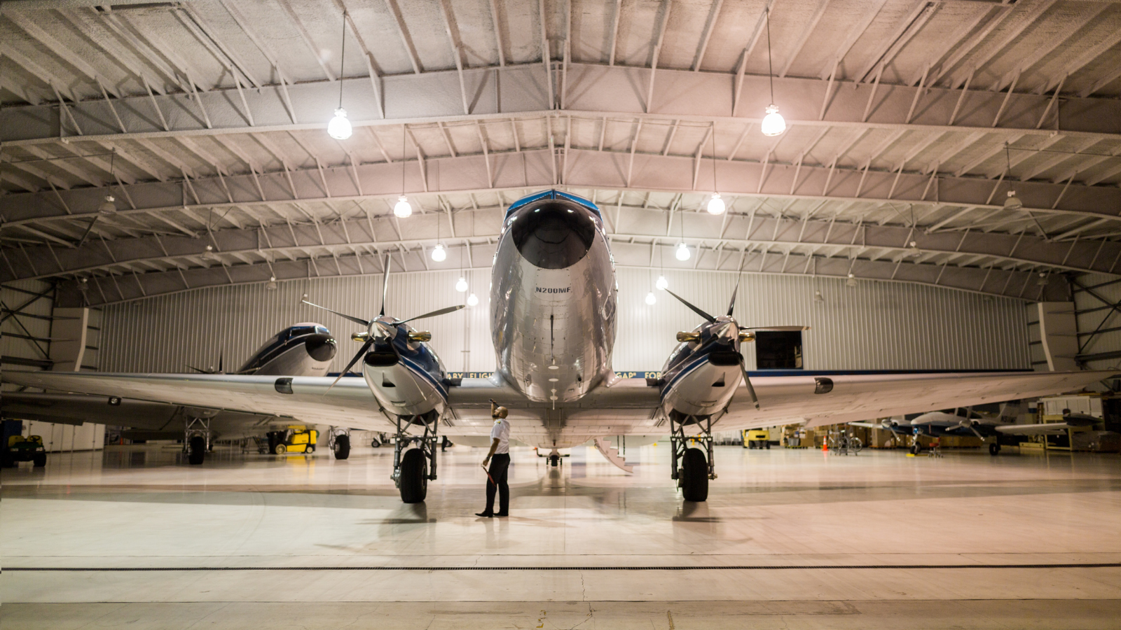 A large airplane with two engines is parked inside a hangar, viewed from the front as a person stands on the floor beneath the aircraft under bright overhead lights.