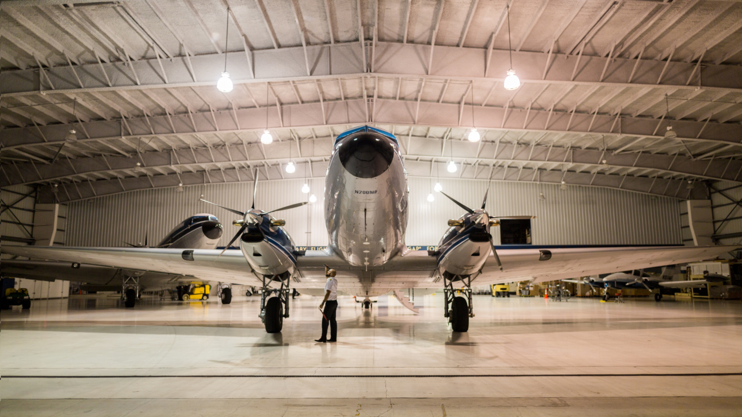 A large airplane with two engines is parked inside a hangar, viewed from the front as a person stands on the floor beneath the aircraft under bright overhead lights.