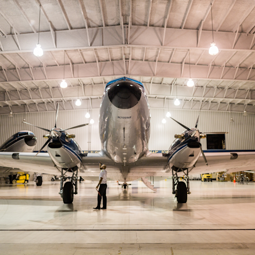 A large airplane with two engines is parked inside a hangar, viewed from the front as a person stands on the floor beneath the aircraft under bright overhead lights.