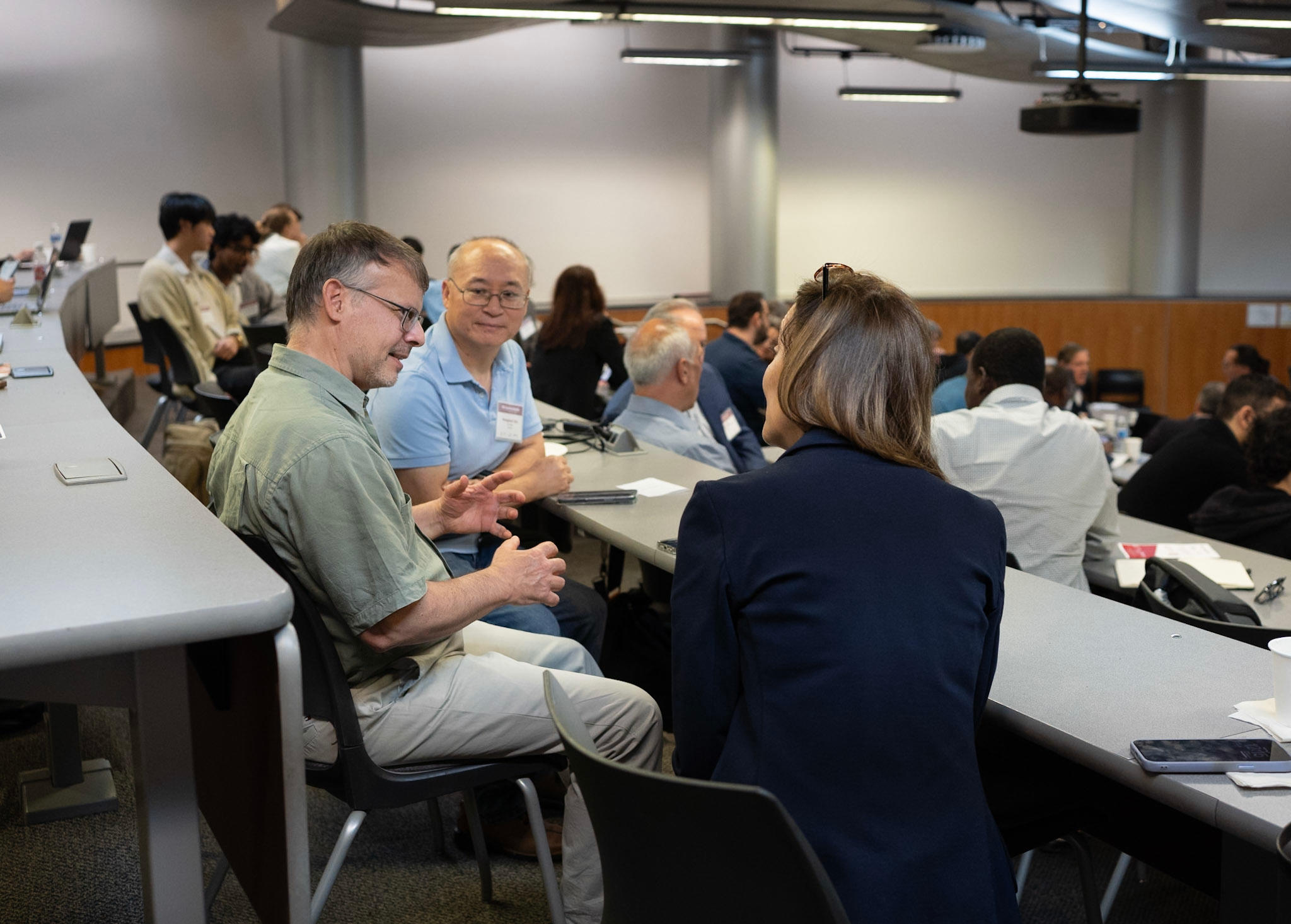 Three adults sit around each other in the same row in a lecture hall discussing the subject at hand.